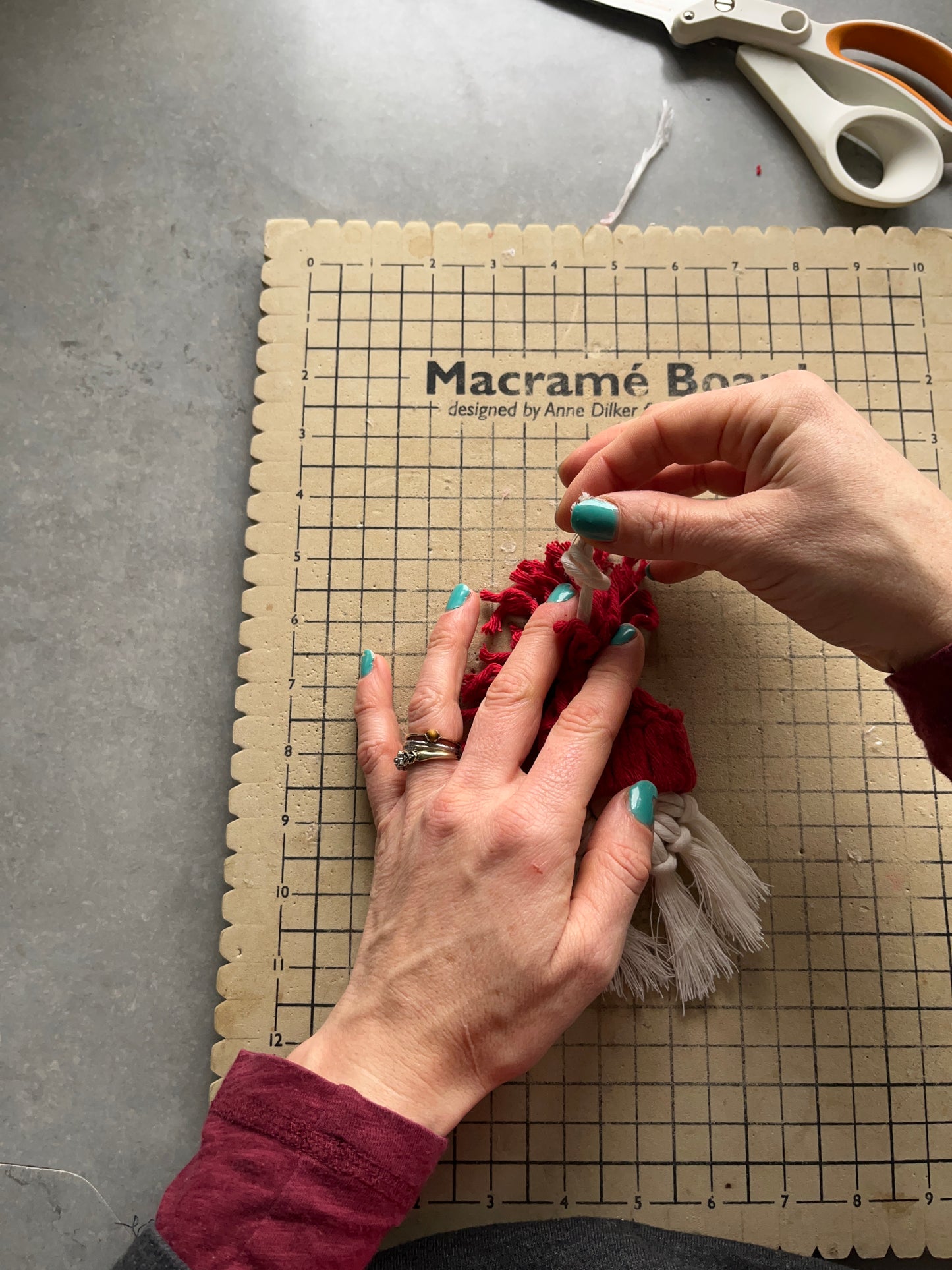 A person's hands are shown crafting a macrame Santa decoration on a macrame board, with cotton cords in red and white.