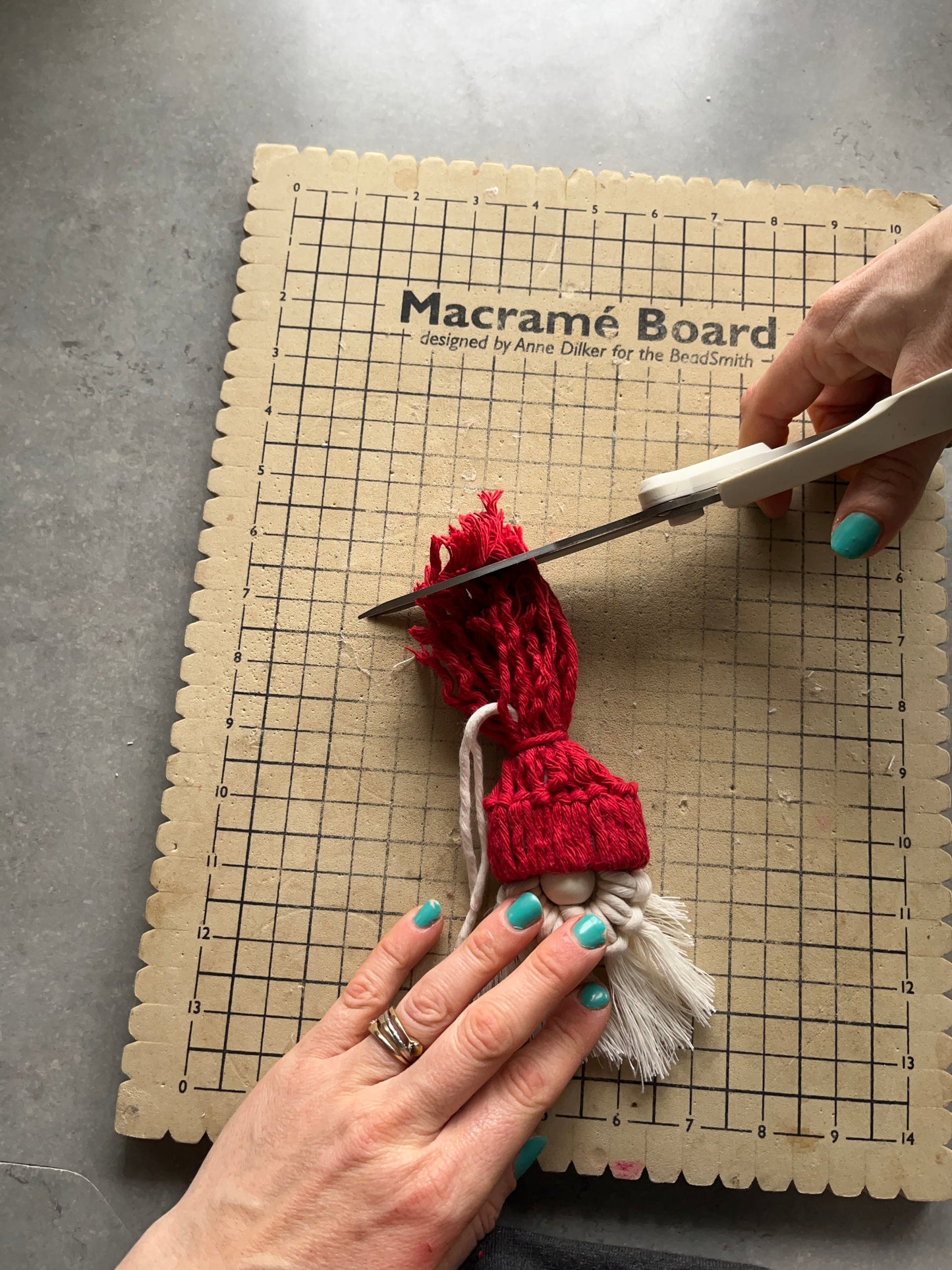 A person's hands are shown crafting a macrame Santa decoration on a macrame board, with cotton cords and wooden bead.