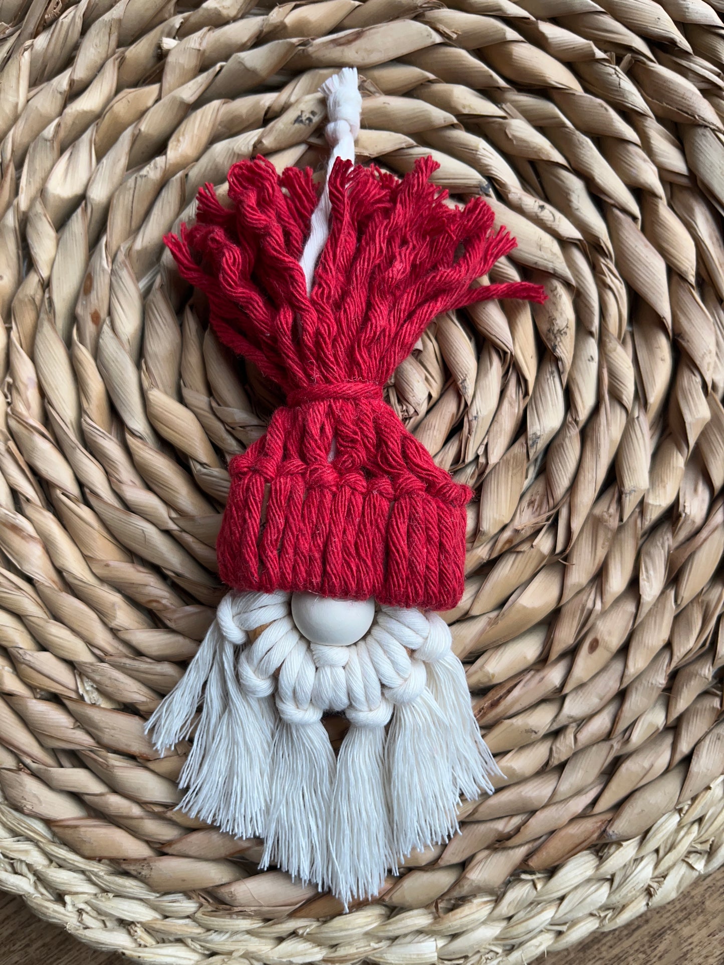A handmade macrame santa hanging decoration made with red and white yarns and a small wooden bead, laid on a round wicker mat.