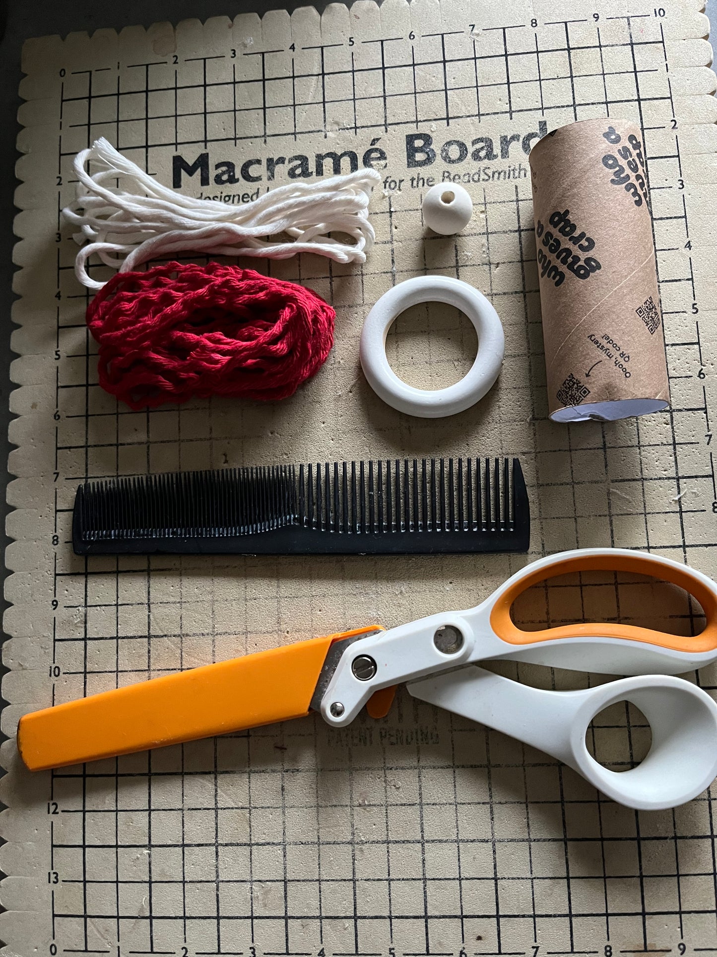 A selection of craft supplies including yarns, wooden ring, wooden bead, toilet roll middle, comb and scissors laid neatly out on a checkered macrame board.
