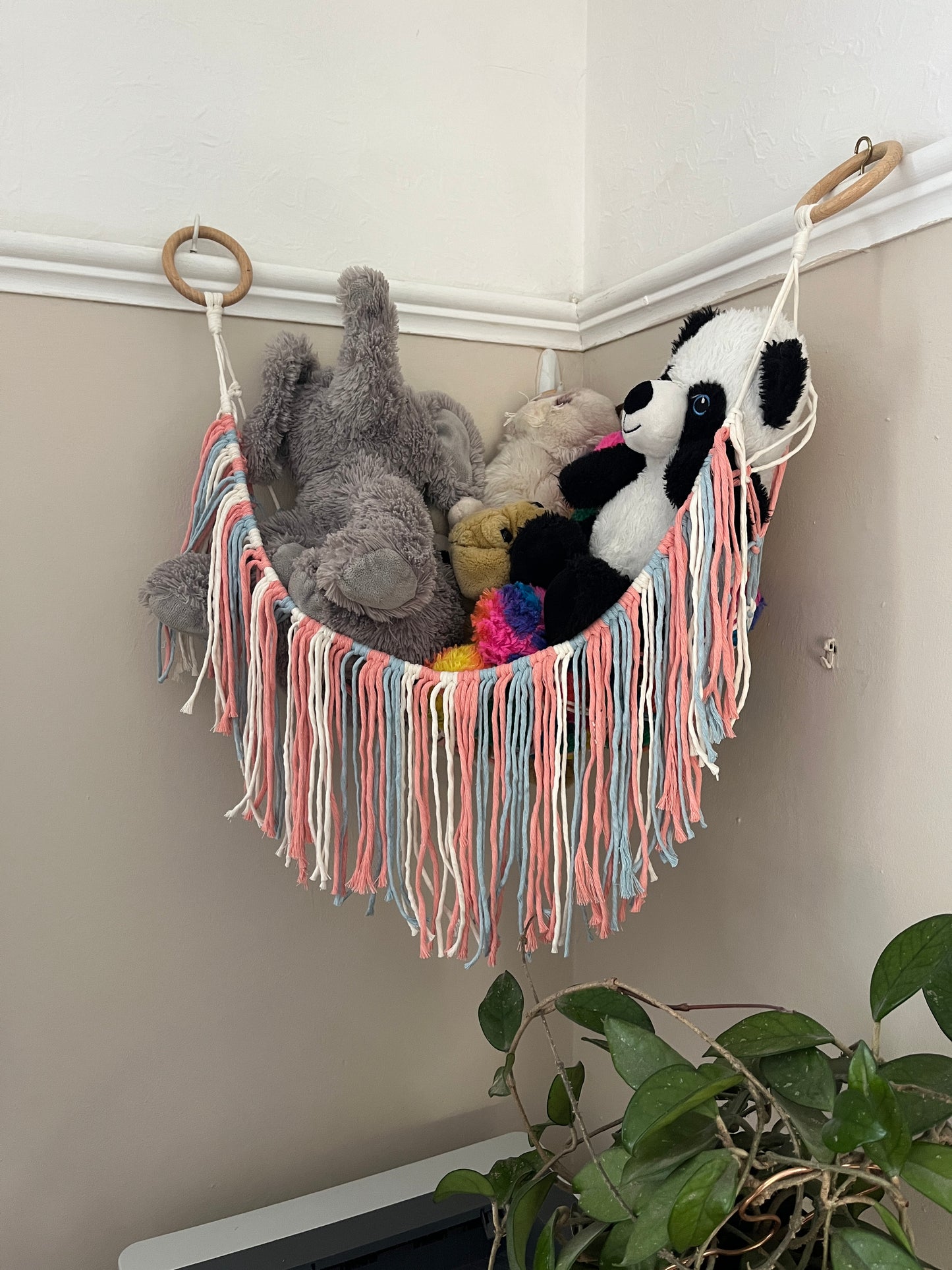 Macrame hammock, made with pink, blue and white yarns filled with colourful soft toys, hanging in the corner of a room with beige painted walls. There is a houseplant below.