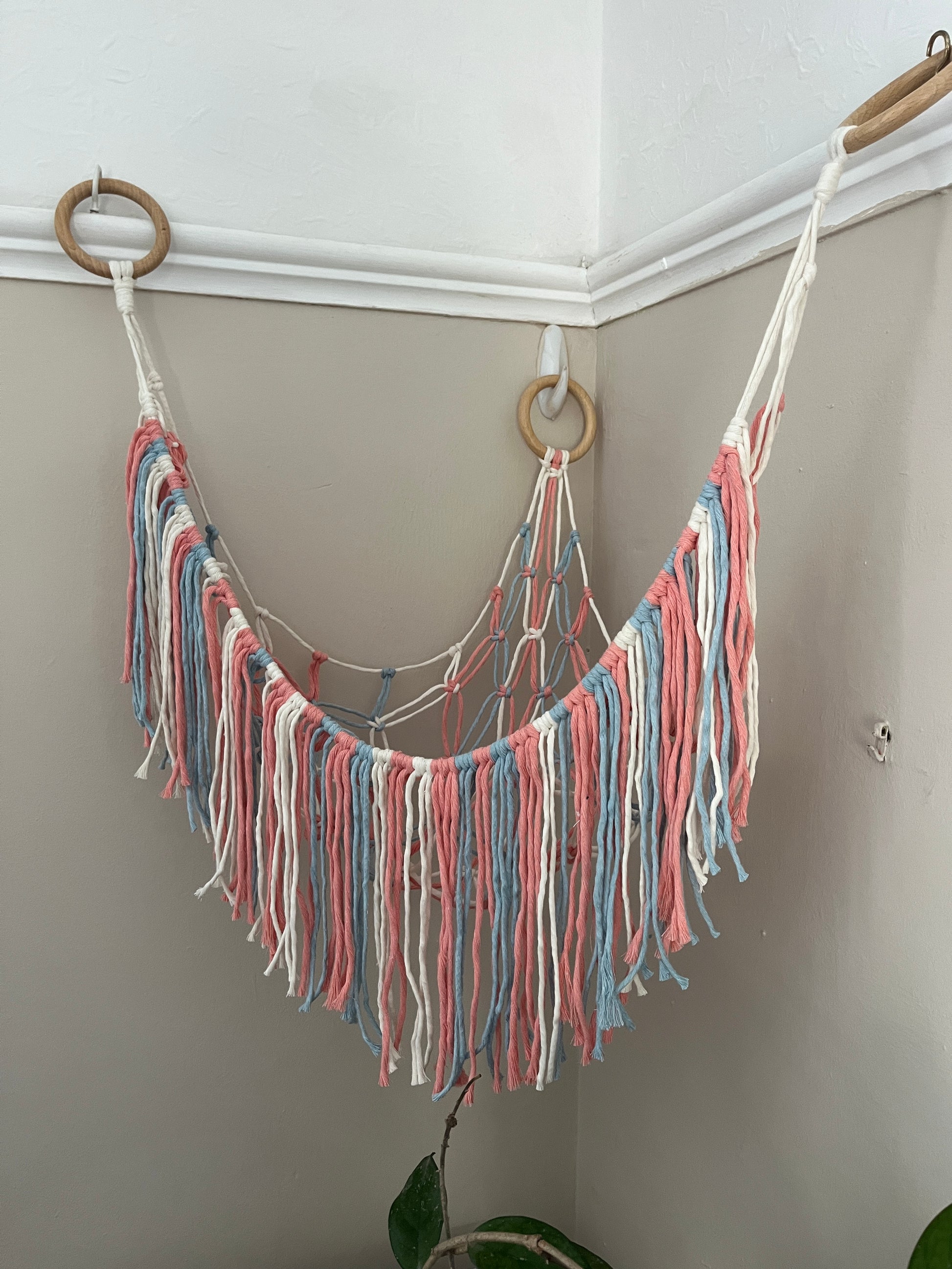Macrame hammock, made with pink, blue and white yarns and three large wooden rings, hanging in the corner of a room with beige painted walls. The hammock is empty.