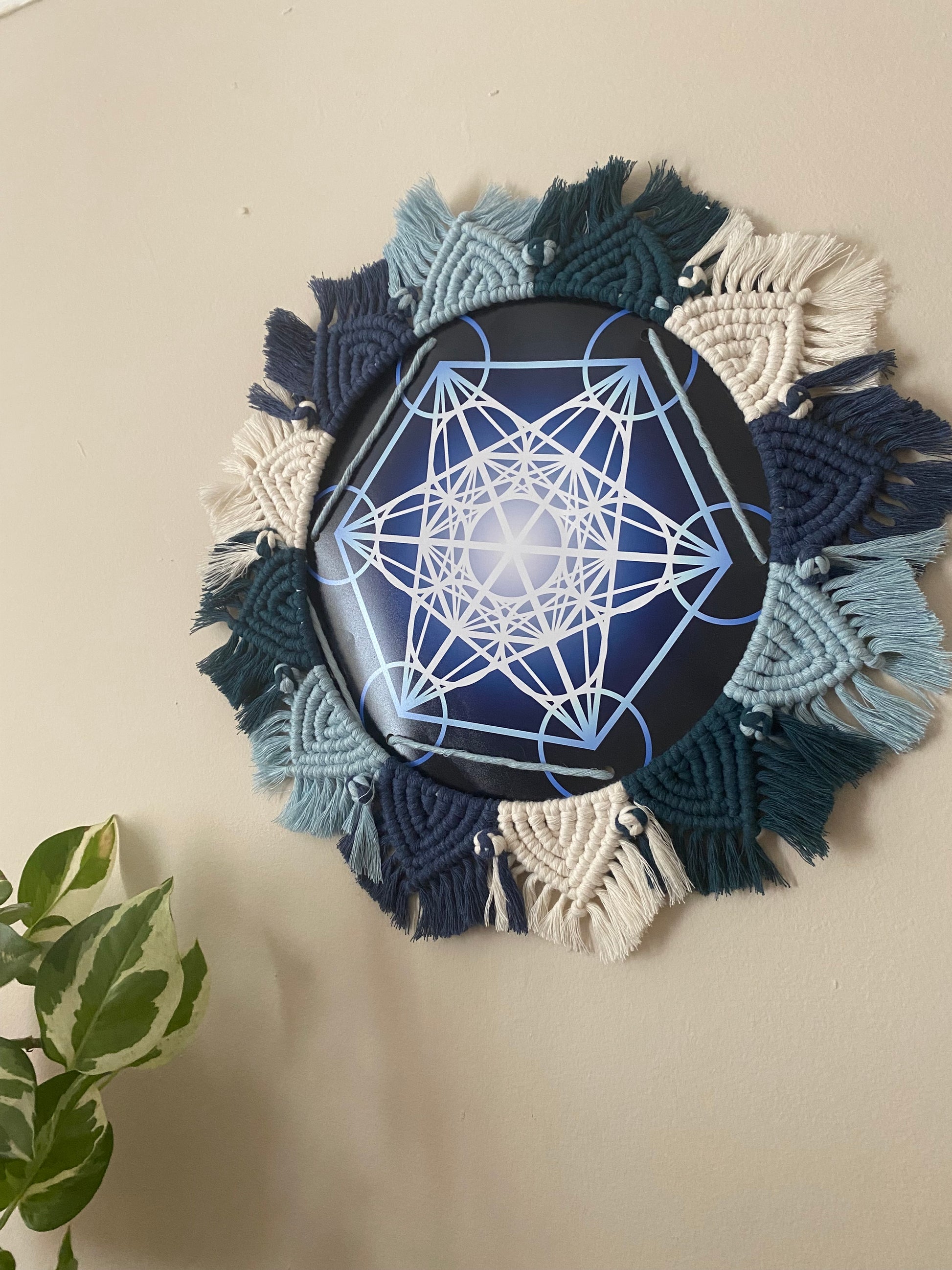A macrame mandala frame knotted with blue and white yarn, surrounds a Metatron’s Cube geometric print. Hung on a beige wall.