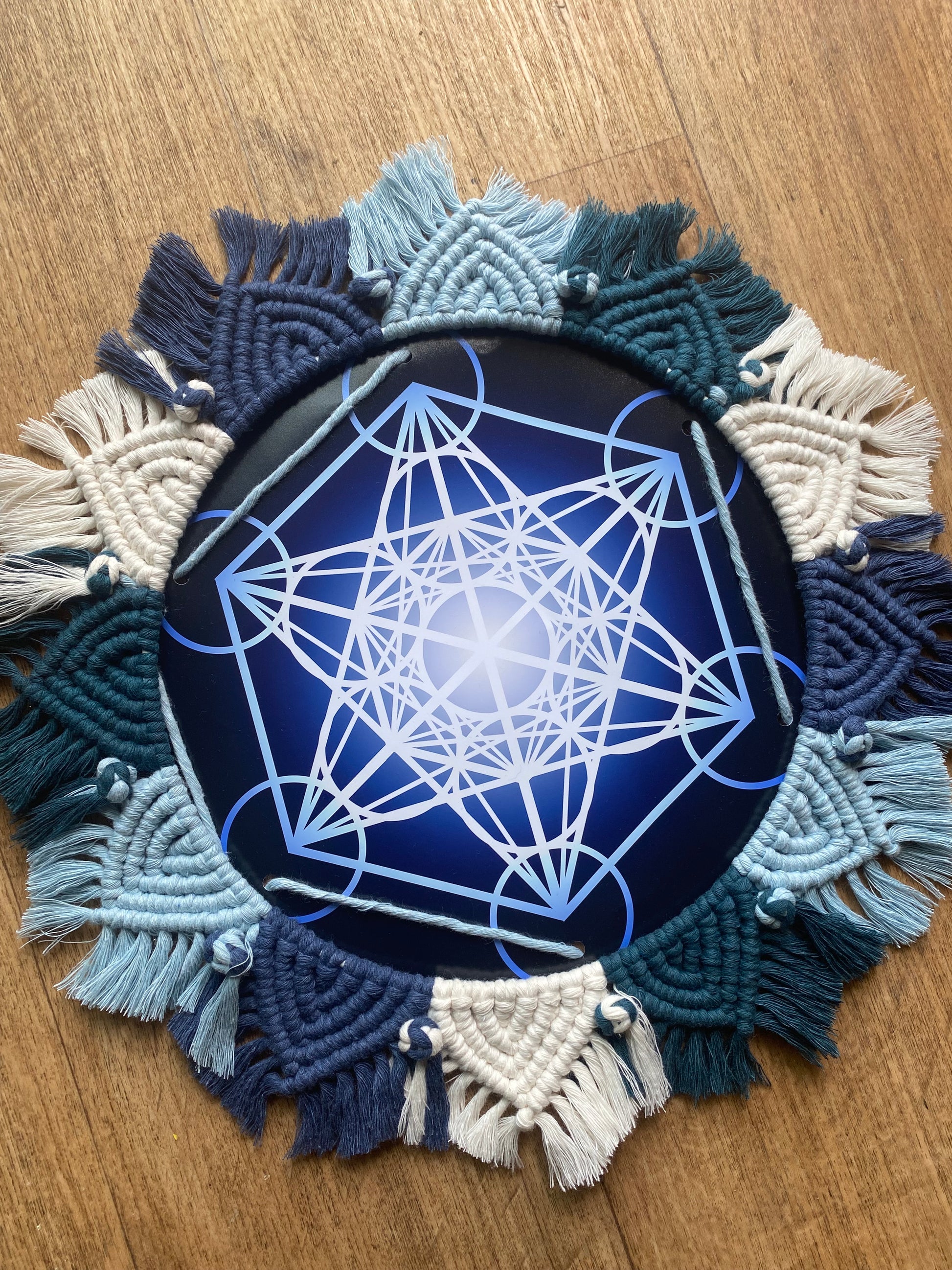 A macrame mandala frame knotted with blue and white yarn, surrounds a Metatron’s Cube geometric print. Laid flat on a wooden floor.