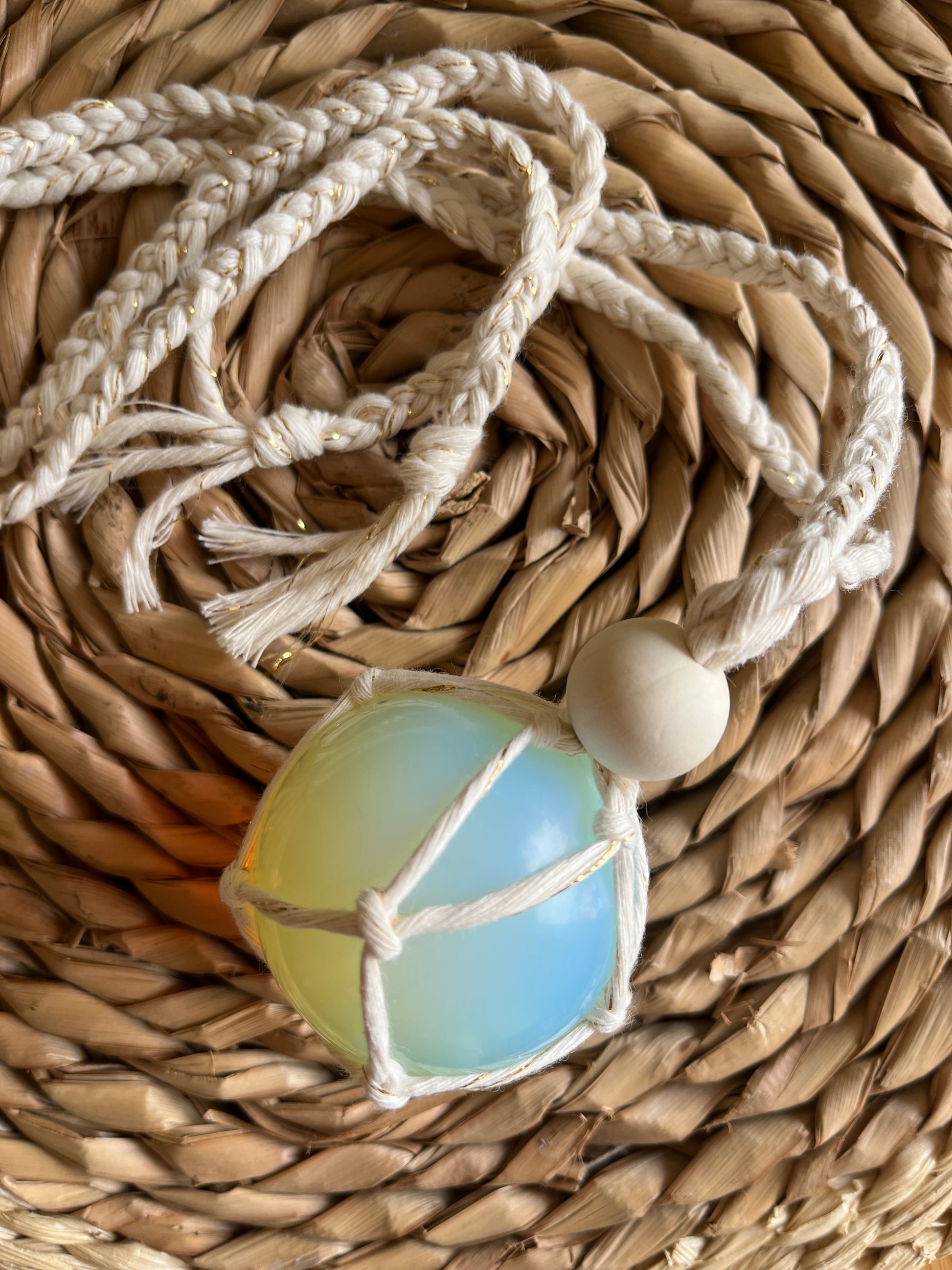 Handmade macrame opaline sphere hanging sun catcher, made with eco friendly natural recycled cotton yarn. The sphere is shown on a wicker surface, there is a plain wooden bead above the crystal.