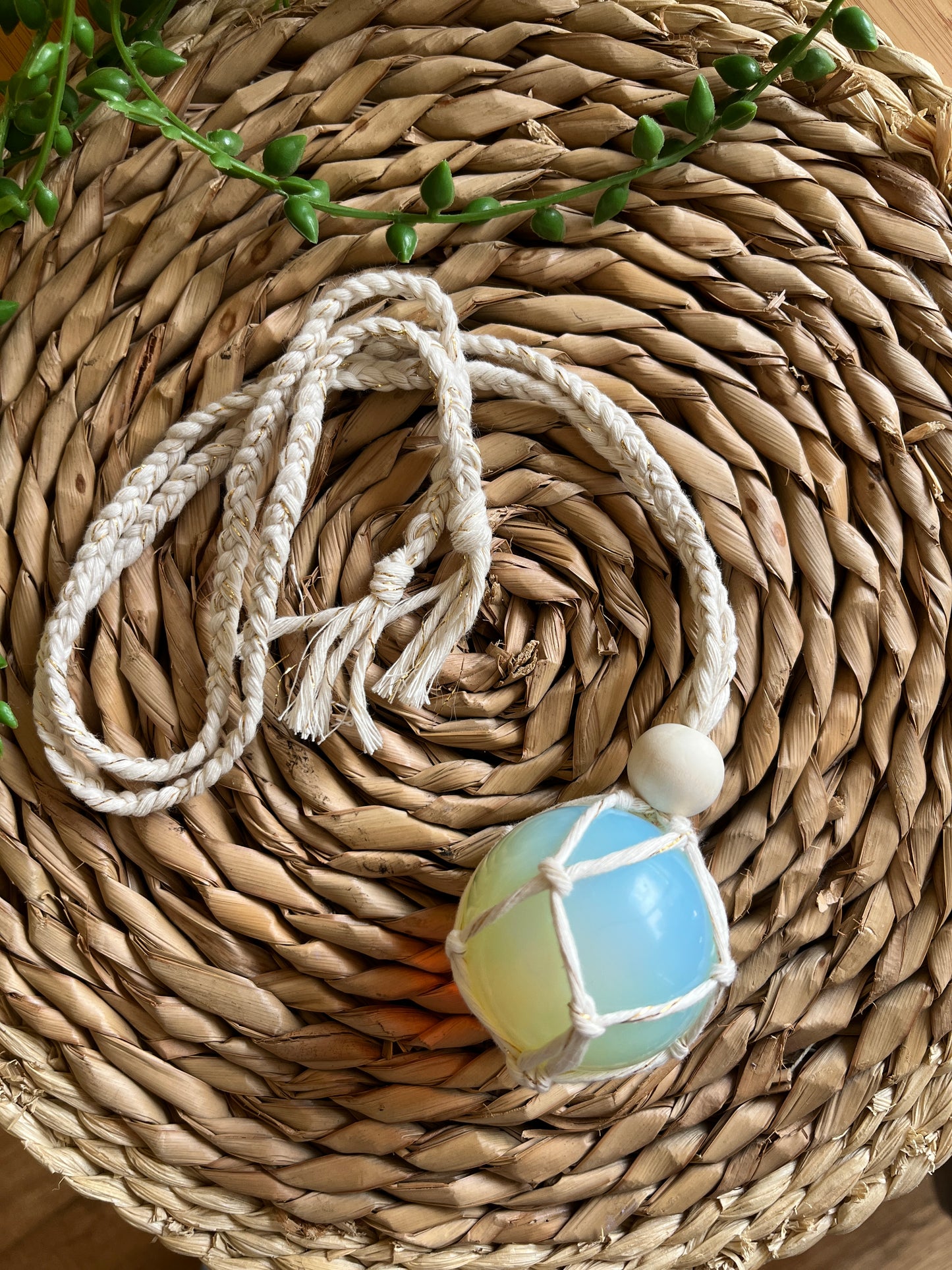 Handmade macrame opaline sphere hanging sun catcher, made with eco friendly natural recycled cotton yarn. The sphere is shown on a wicker surface, there is a plain wooden bead above the crystal.
