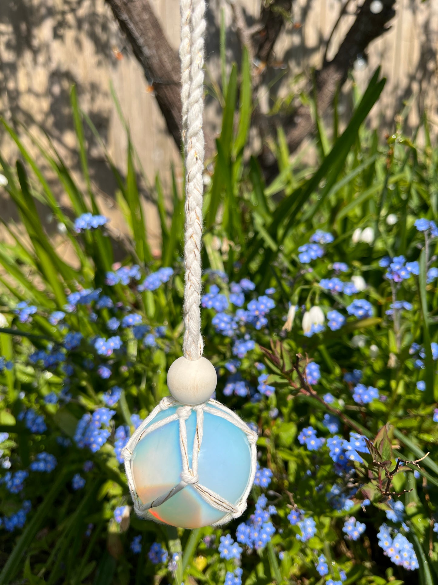 Handmade macrame opaline sphere hanging sun catcher, made with eco friendly natural recycled cotton yarn. The sphere is shown hanging outside in front of some small blue flowers, there is a plain wooden bead above the crystal.