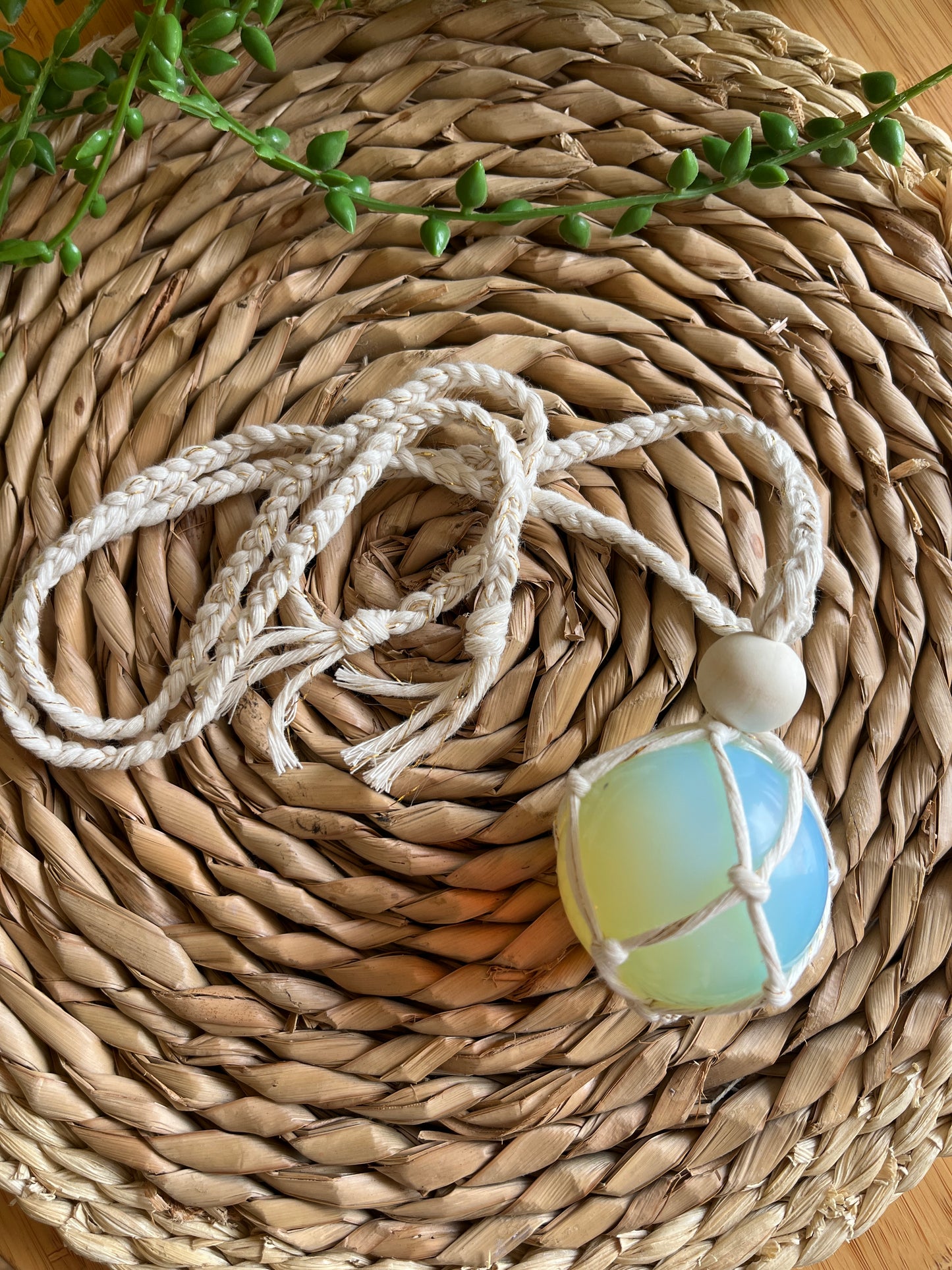 Handmade macrame opaline sphere hanging sun catcher, made with eco friendly natural recycled cotton yarn. The sphere is shown on a wicker surface, there is a plain wooden bead above the crystal.