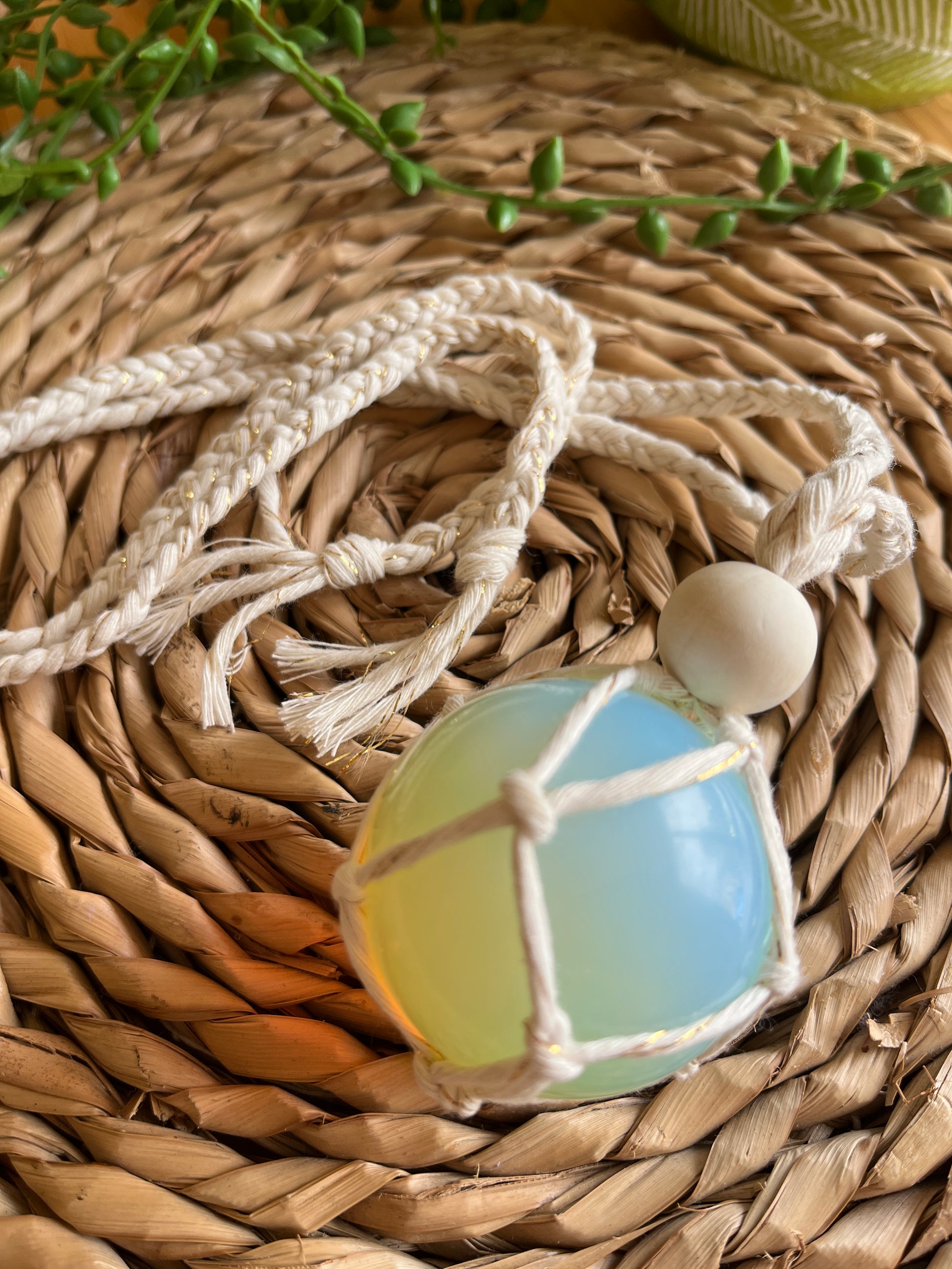 Handmade macrame opaline sphere hanging sun catcher, made with eco friendly natural recycled cotton yarn. The sphere is shown on a wicker surface, there is a plain wooden bead above the crystal.