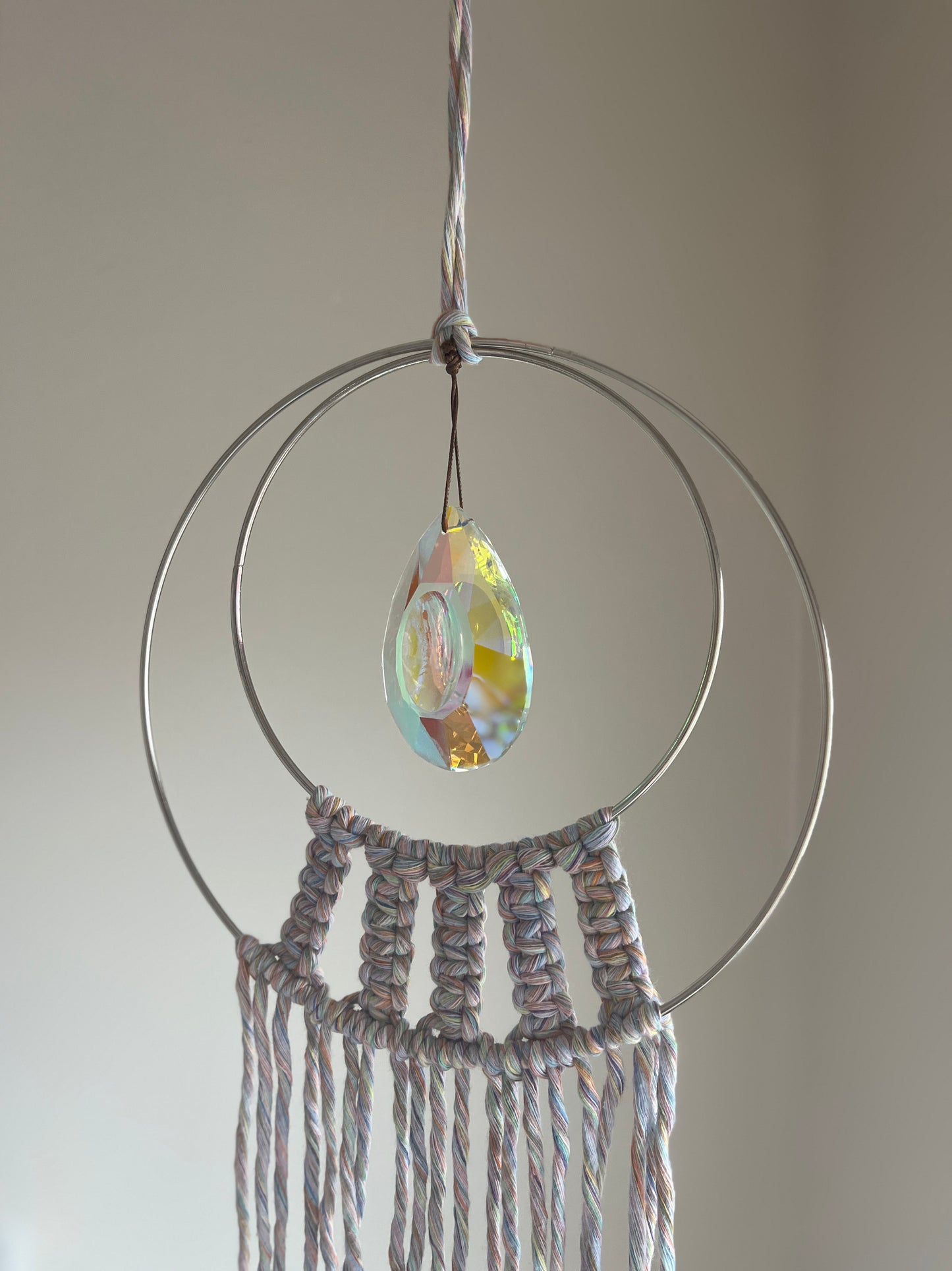 Handmade macrame Suncatcher made with pastel coloured recycled yarn, on two metal hoops, with a large hanging crystal in the centre, shown hanging in front of a white wall.