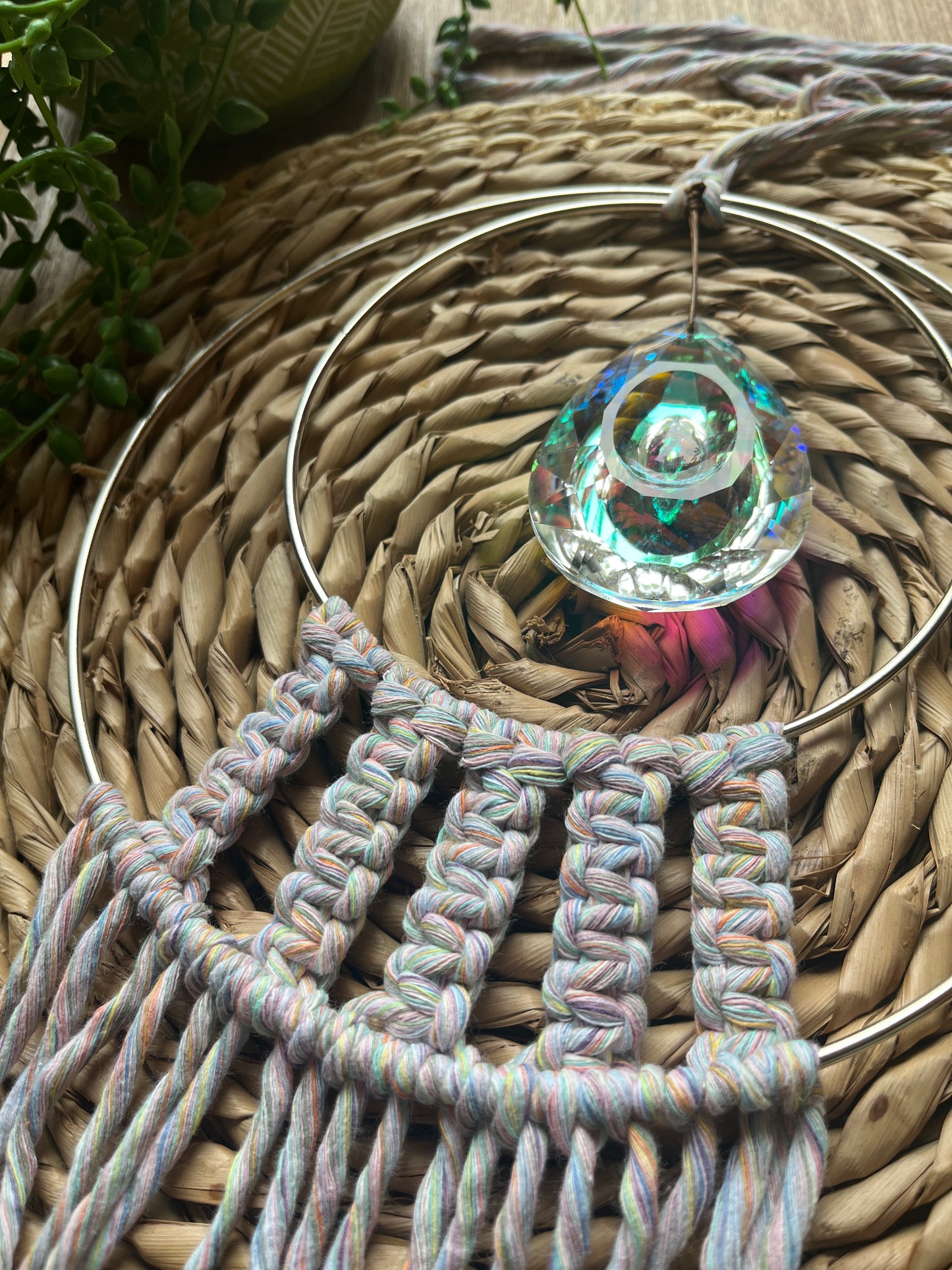 Handmade macrame Suncatcher made with pastel coloured recycled yarn, on two metal hoops, with a large hanging crystal in the centre, on a wicker surface in the sunshine.