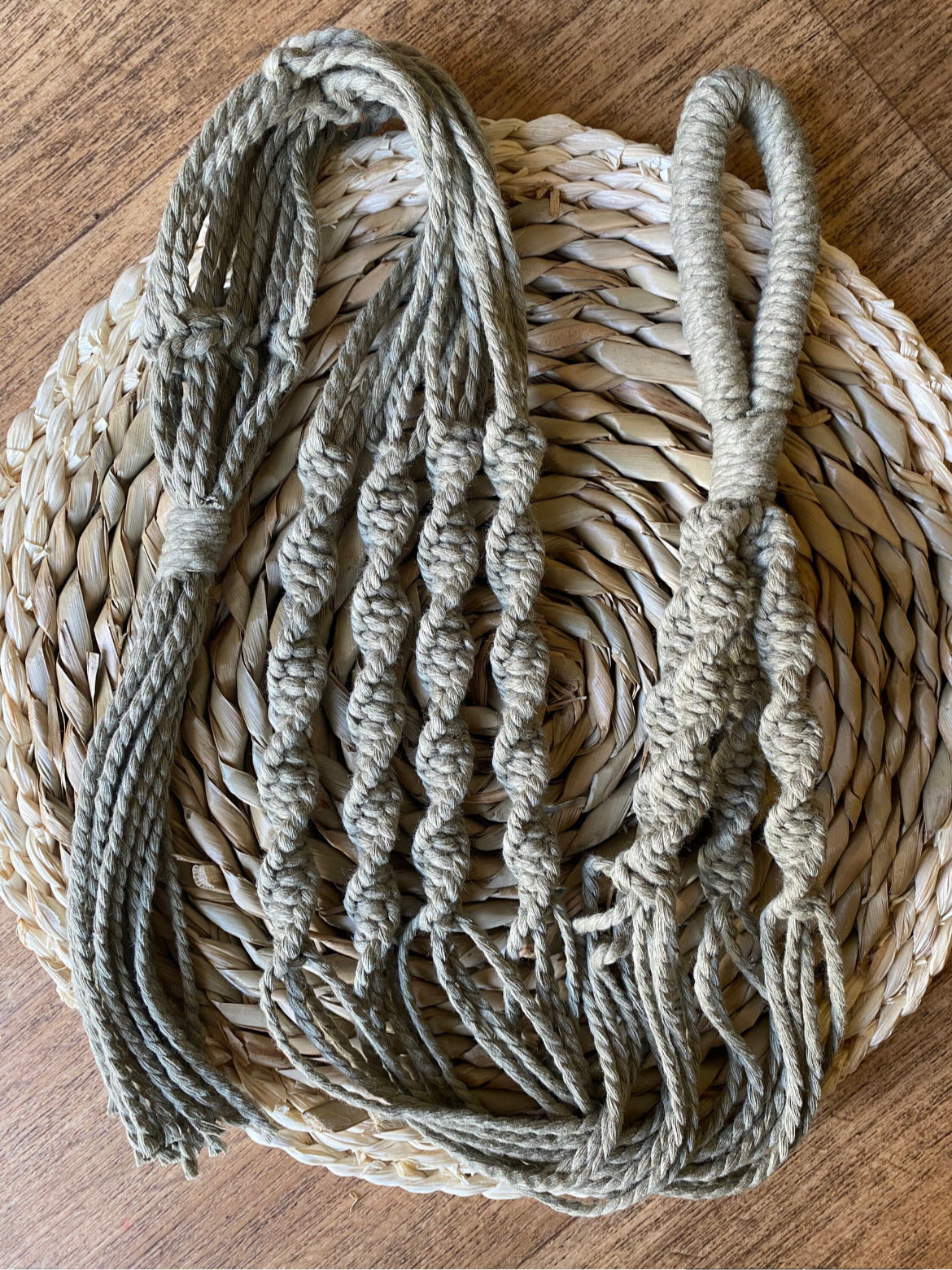 Handmade Macrame plant hanger, made with eco friendly sage green recycled cotton. The plant hanger is laid out on a woven surface.