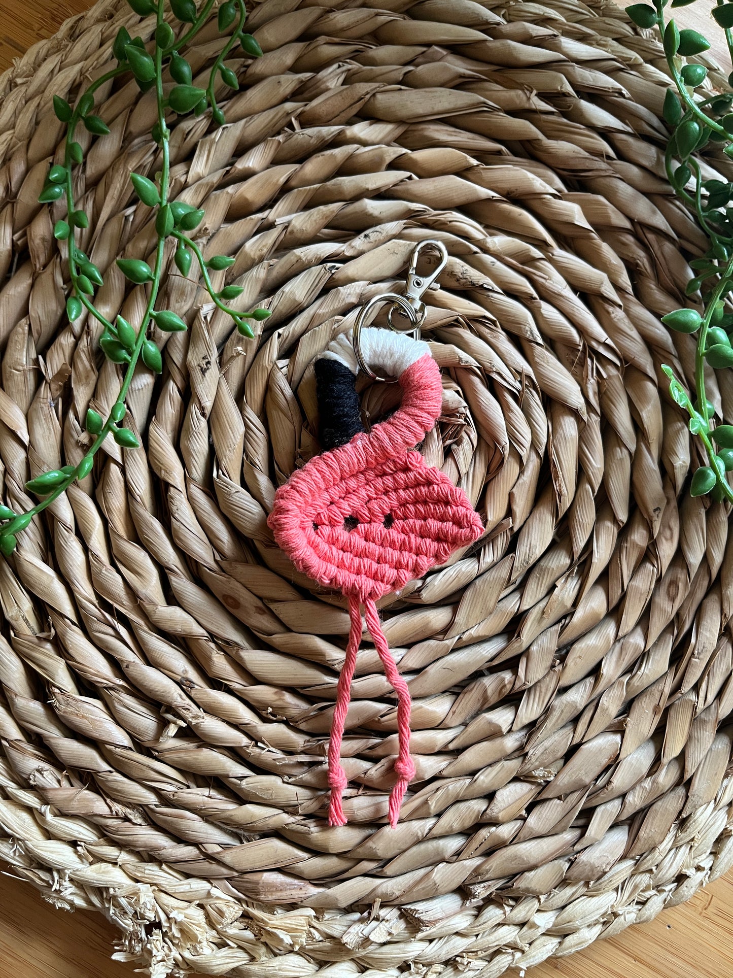 Handmade macrame keychain in the shape of a flamingo, made using salmon pink recycled cotton, on a metal split ring with lobster clasp. The keyring is displayed on a round wicker surface next to some green plant vines.