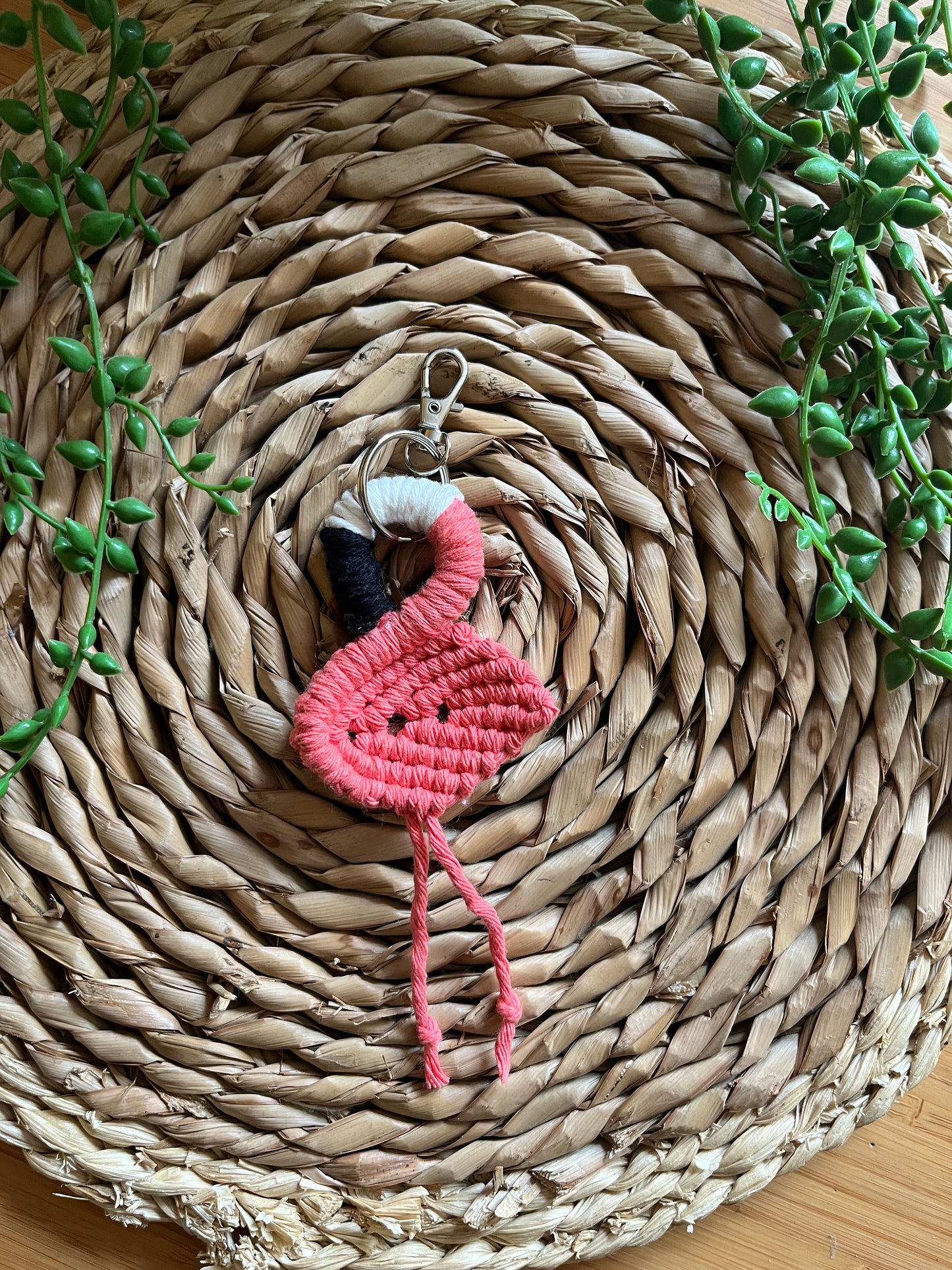Handmade macrame keychain in the shape of a flamingo, made using salmon pink recycled cotton, on a metal split ring with lobster clasp. The keyring is displayed on a round wicker surface next to some green plant vines.