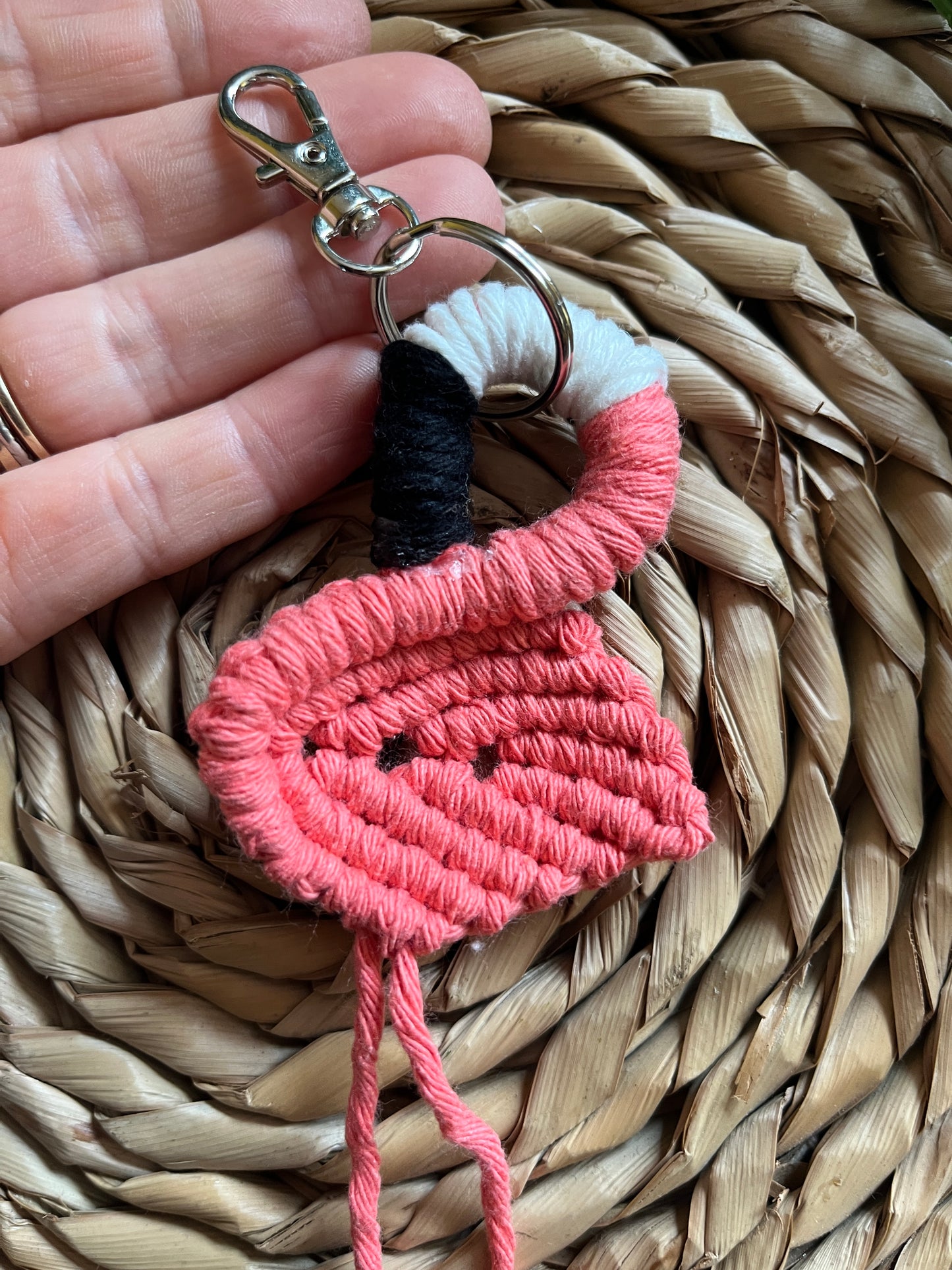 Handmade macrame keychain in the shape of a flamingo, made using salmon pink recycled cotton, on a metal split ring with lobster clasp. The keyring is displayed on a round wicker surface next to some green plant vines.