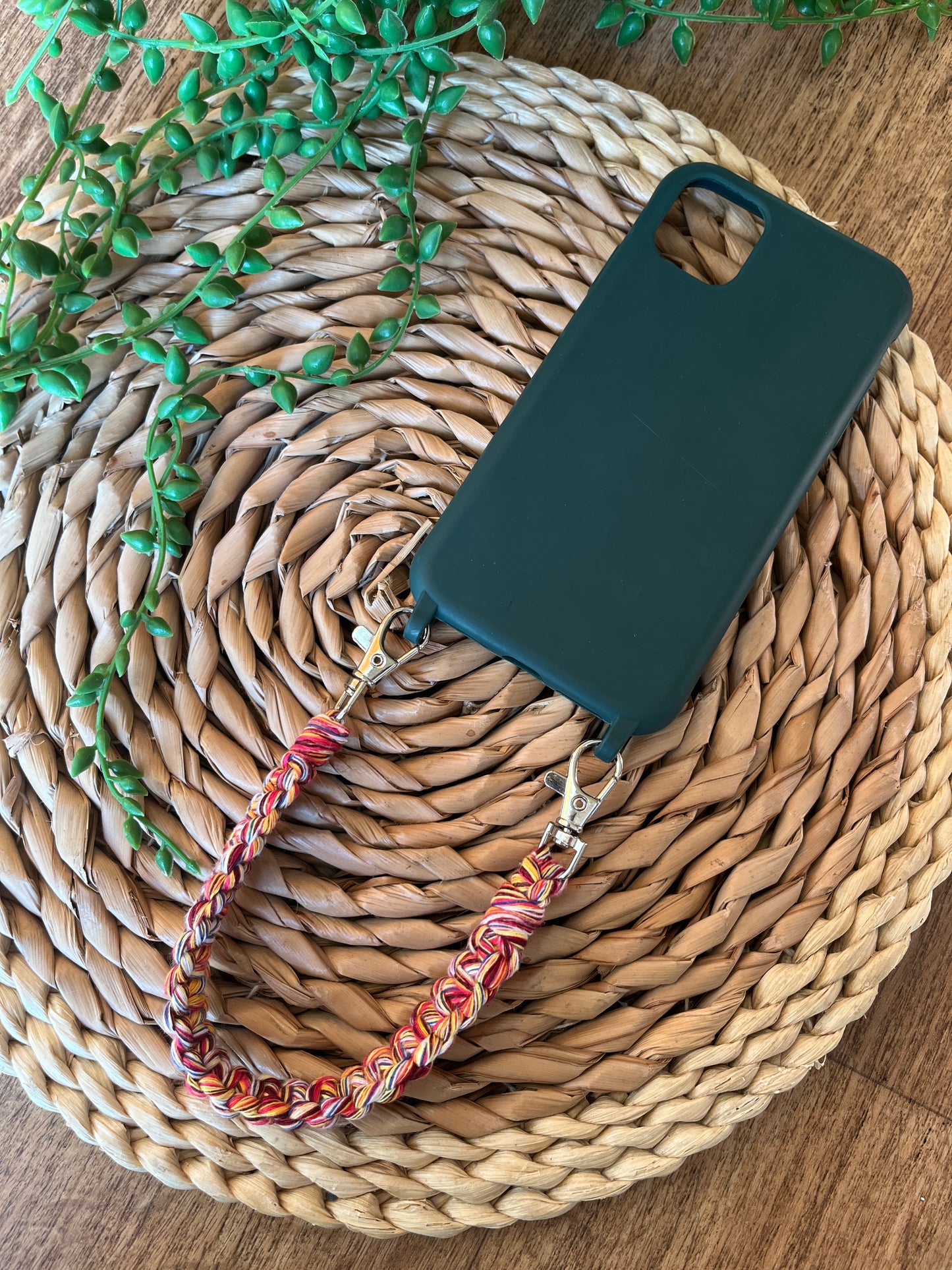 Handmade short macrame phone wristlet loop for phone case. Shown attached to a green phone case on a wicker mat. Handmade with multicoloured recycled cotton yarn, with a small metal lobster clasp on each end.