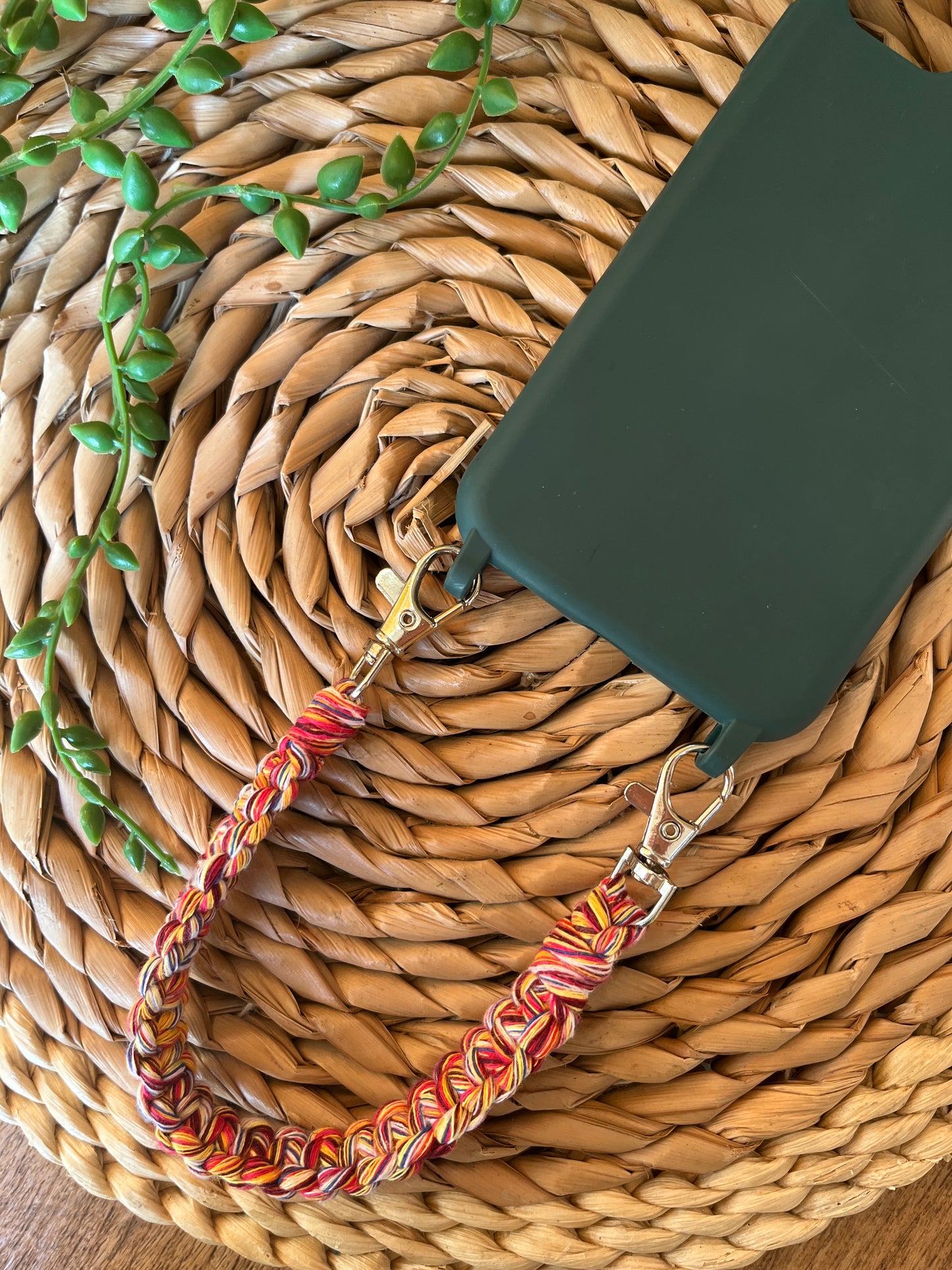 Handmade short macrame phone wristlet loop for phone case. Shown attached to a green phone case on a wicker mat. Handmade with multicoloured recycled cotton yarn, with a small metal lobster clasp on each end.