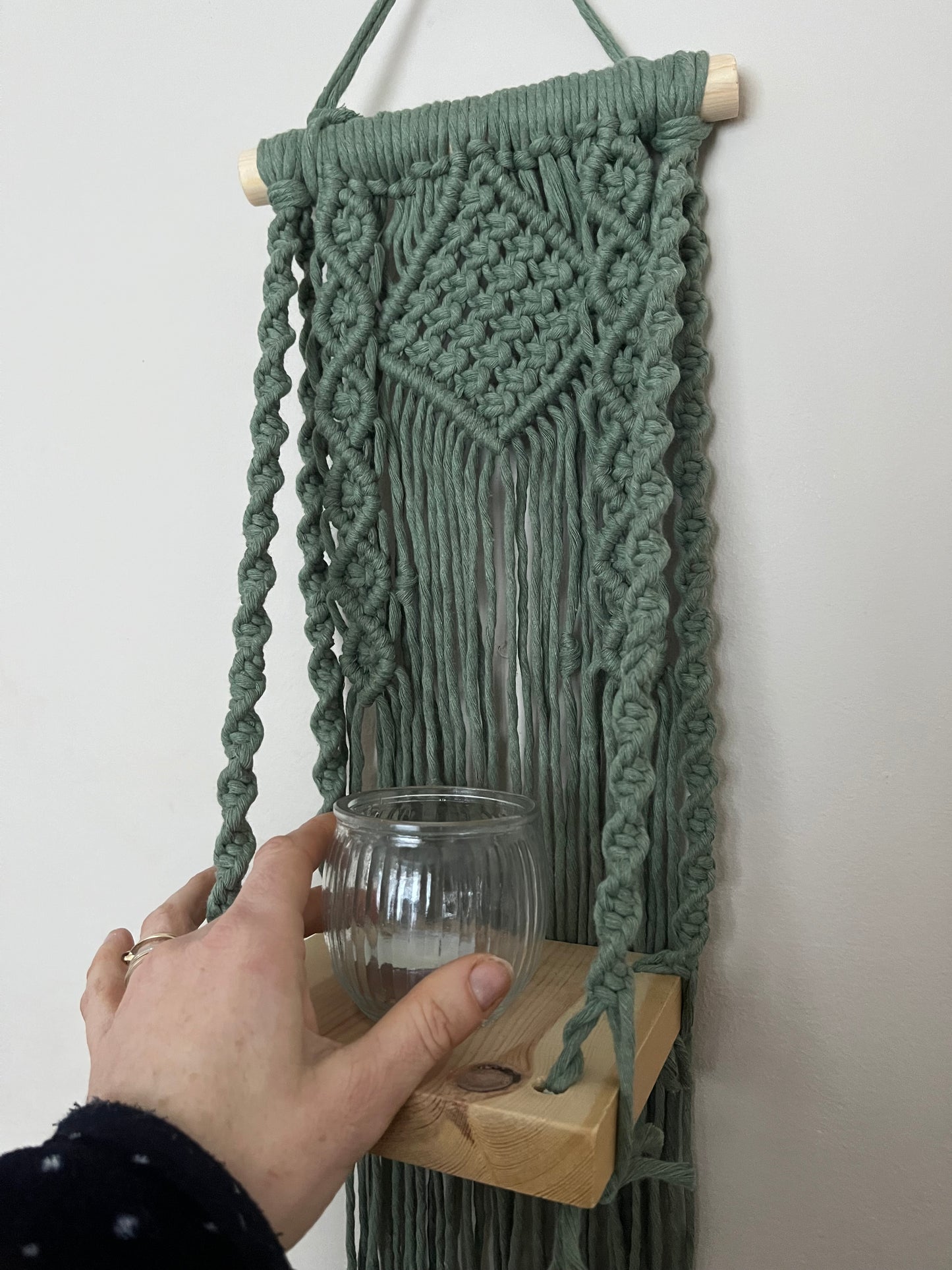 Handmade macrame boho style shelf, made with sage green recycled cotton on a solid wooden dowel, with a small square shelf. The shelf has an intricately knotted pattern. A hand is seen, placing a small glass candle jar on to the shelf.
