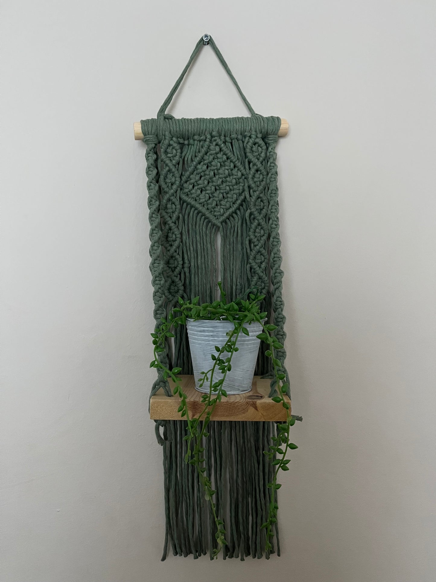 Handmade macrame boho style shelf, made with sage green recycled cotton on a solid wooden dowel, with a small square shelf. The shelf has an intricately knotted pattern, and displays a small artificial plant.