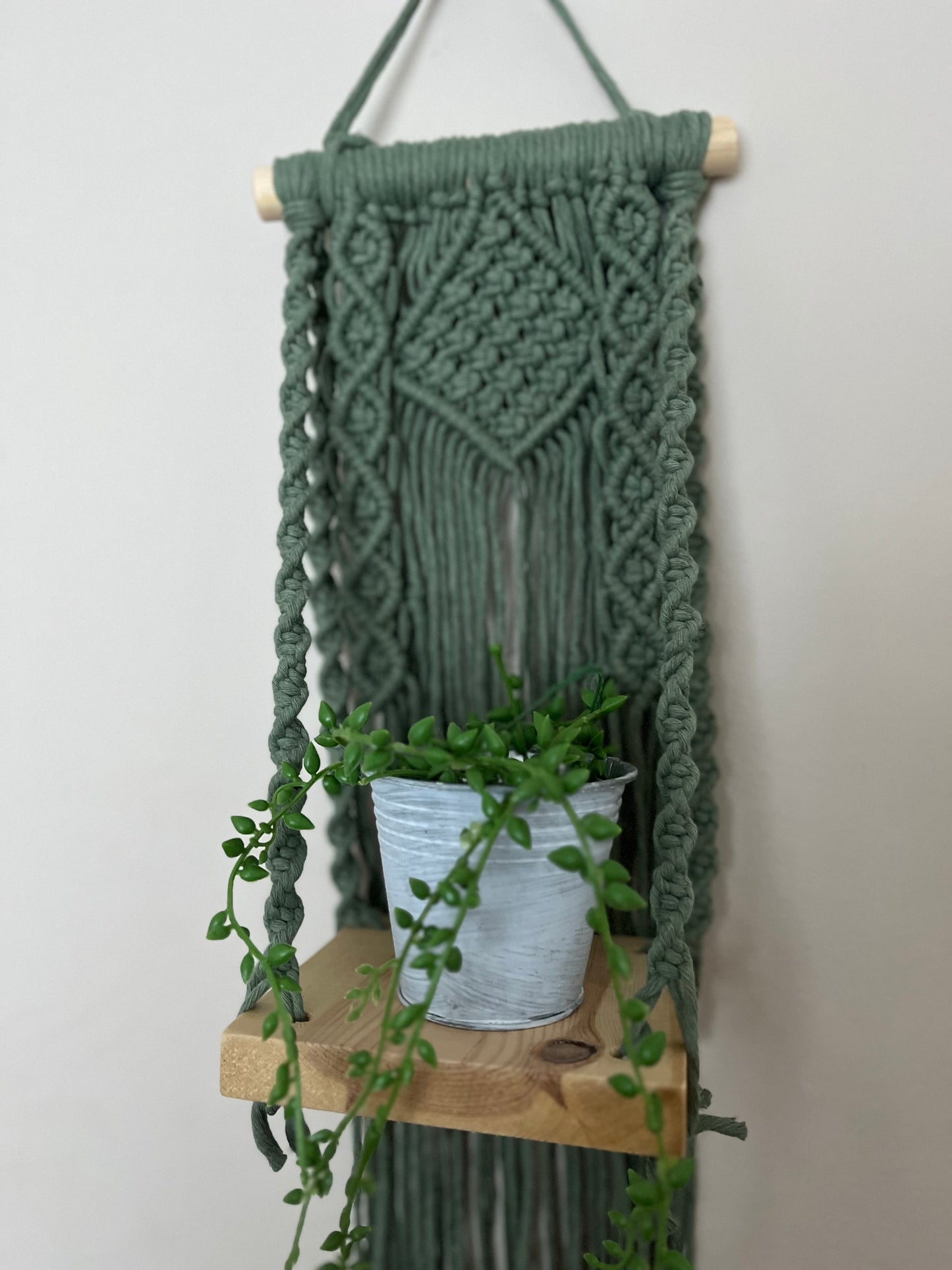 Handmade macrame boho style shelf, made with sage green recycled cotton on a solid wooden dowel, with a small square shelf. The shelf has an intricately knotted pattern, and displays a small artificial plant.