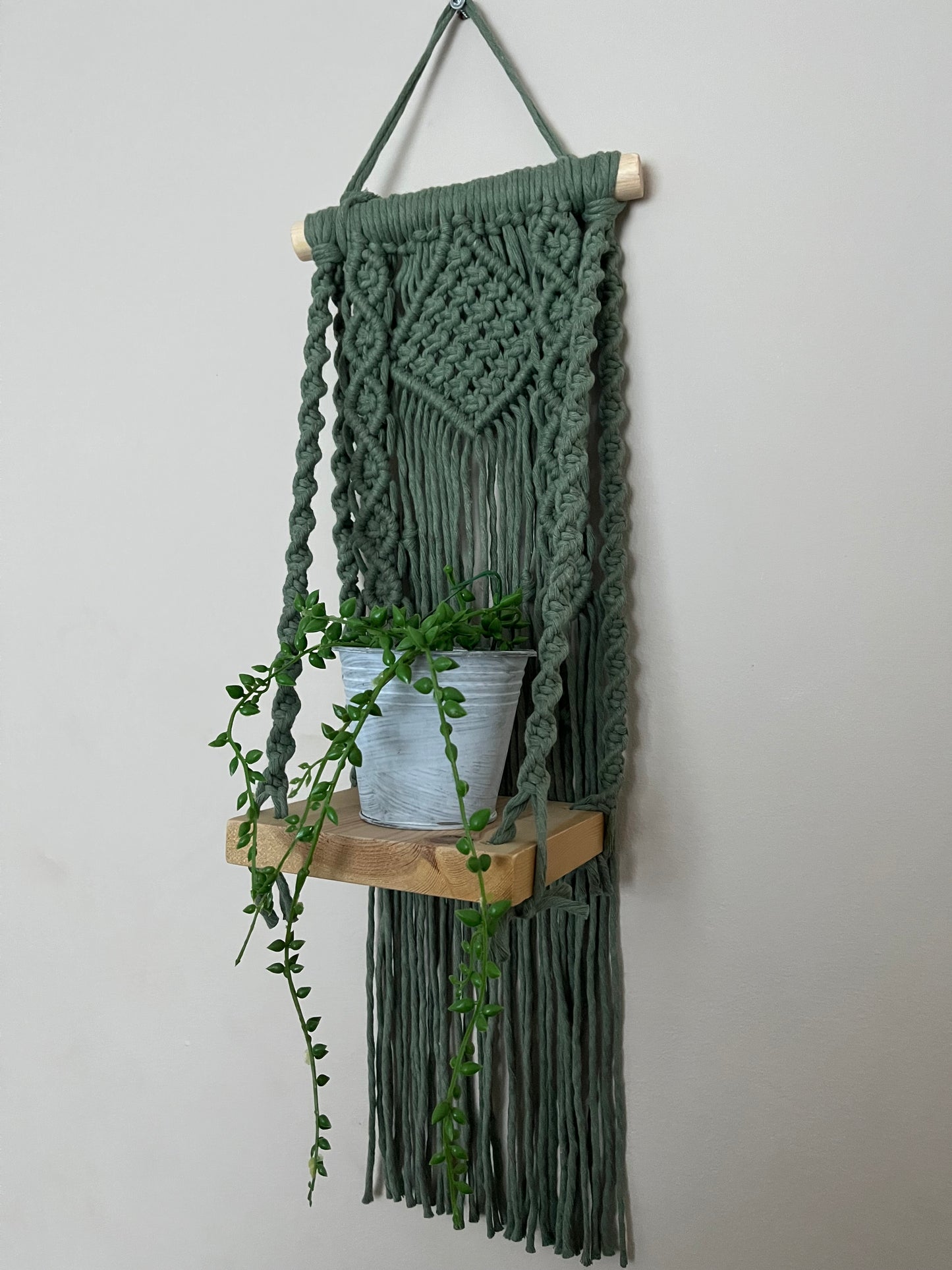 Handmade macrame boho style shelf, made with sage green recycled cotton on a solid wooden dowel, with a small square shelf. The shelf has an intricately knotted pattern, and displays a small artificial plant.