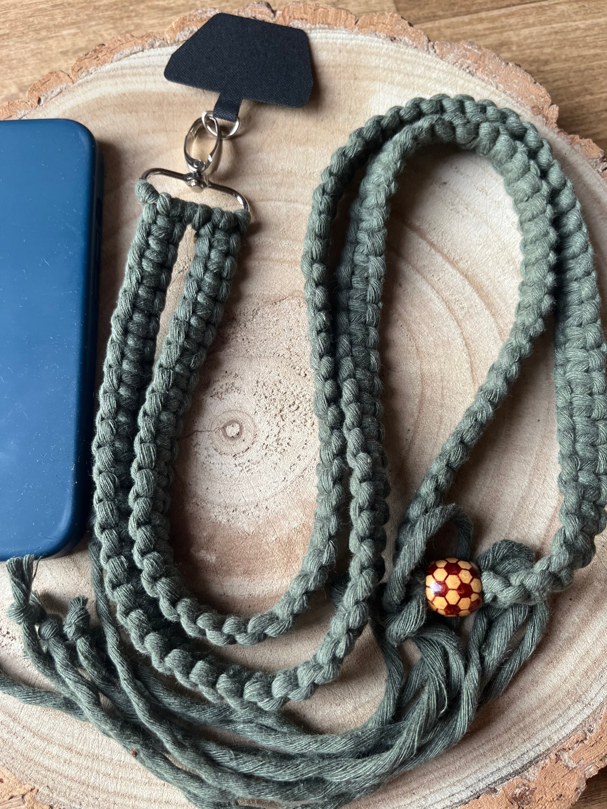 A long crossbody phone strap made with green yarn, a patterned wooden bead and metal clasp has a universal tether attached to it, and is coiled on a round wooden surface next to a blue phone case.