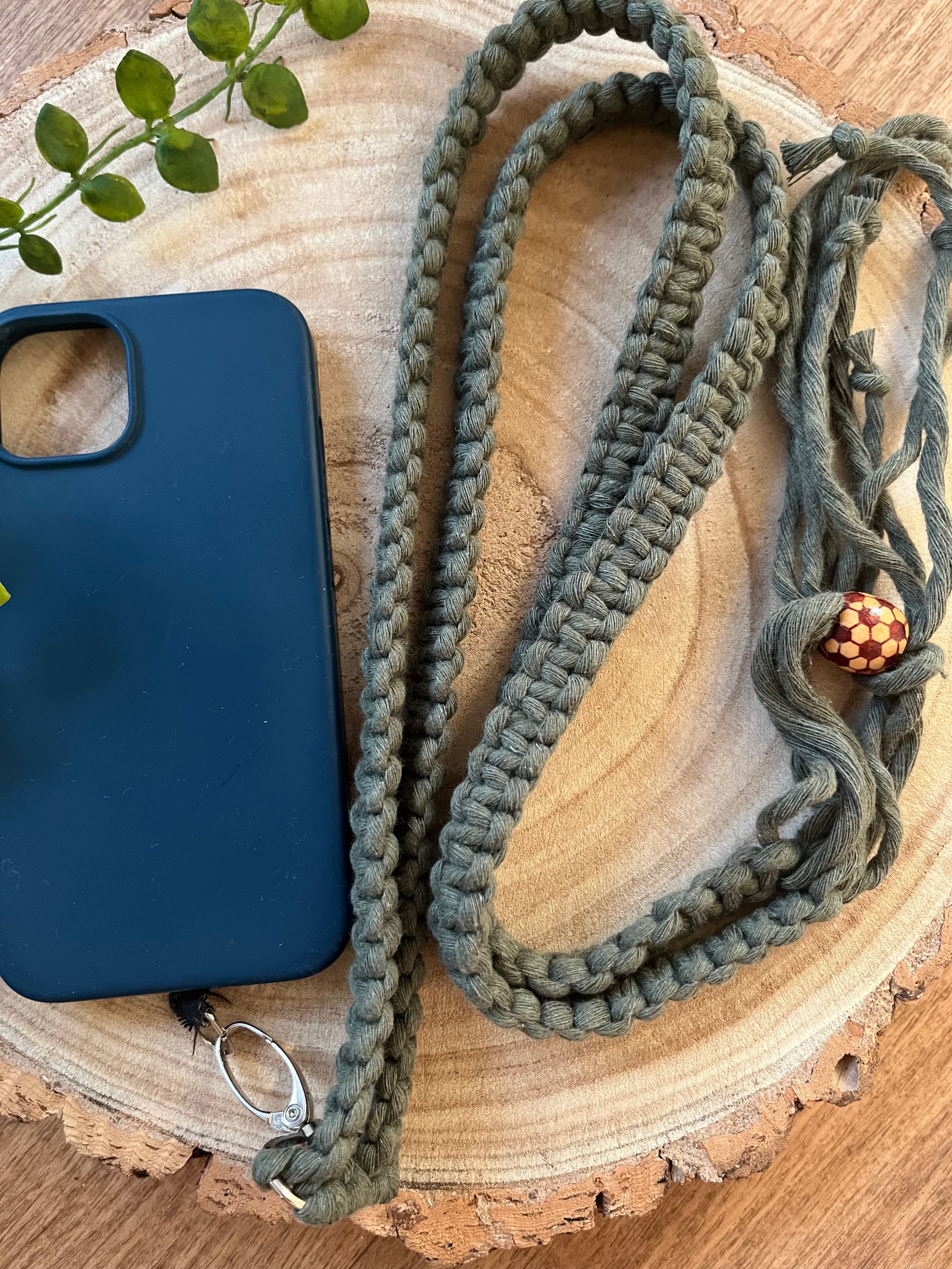 A long crossbody phone strap made with green yarn, a patterned wooden bead and metal clasp is attached to an empty blue phone case, on a round wooden surface.