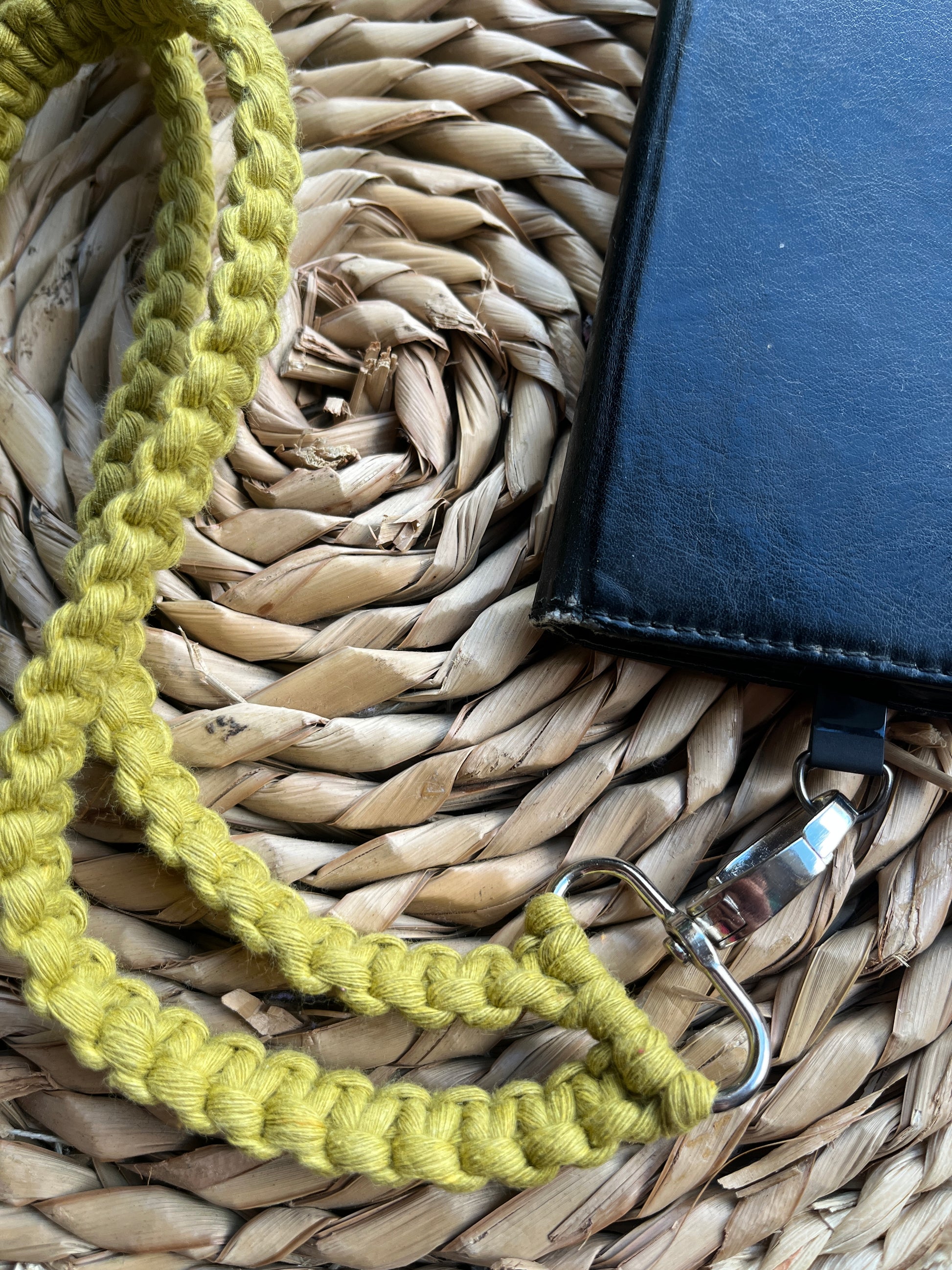Close up of a long crossbody phone strap made with green yarn, attached to a black leather phone case on a round wicker placemat.