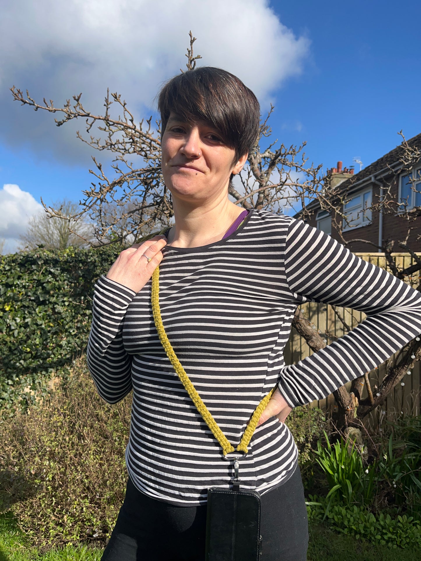 A woman with short brown hair stands outside wearing a striped top. She has a crossbody phone strap across her body, attached to a black phone case.