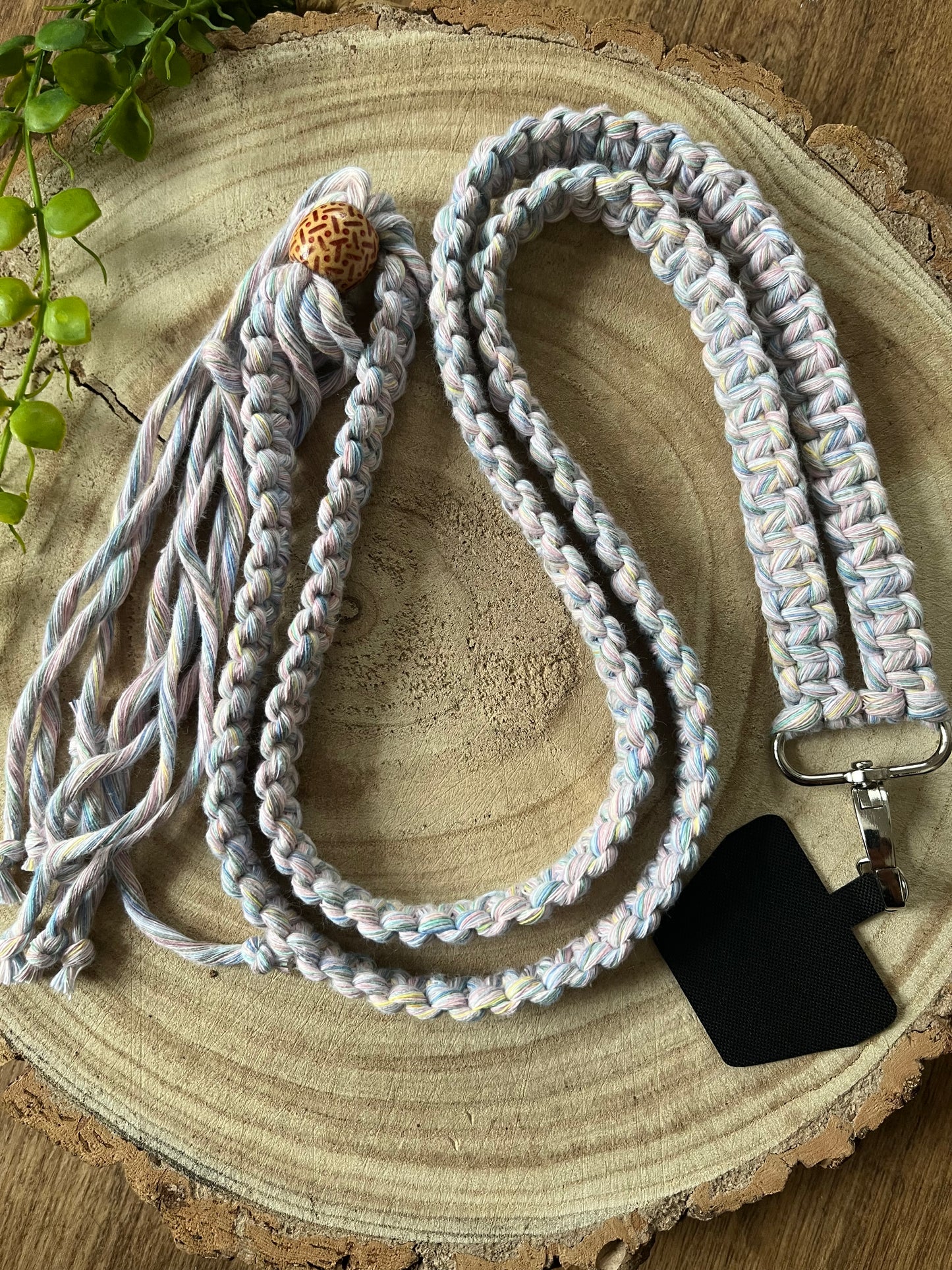 A long crossbody phone strap made with pastel yarn, a patterned wooden bead and metal clasp has a universal tether attached to it, and is coiled on a round wooden surface.