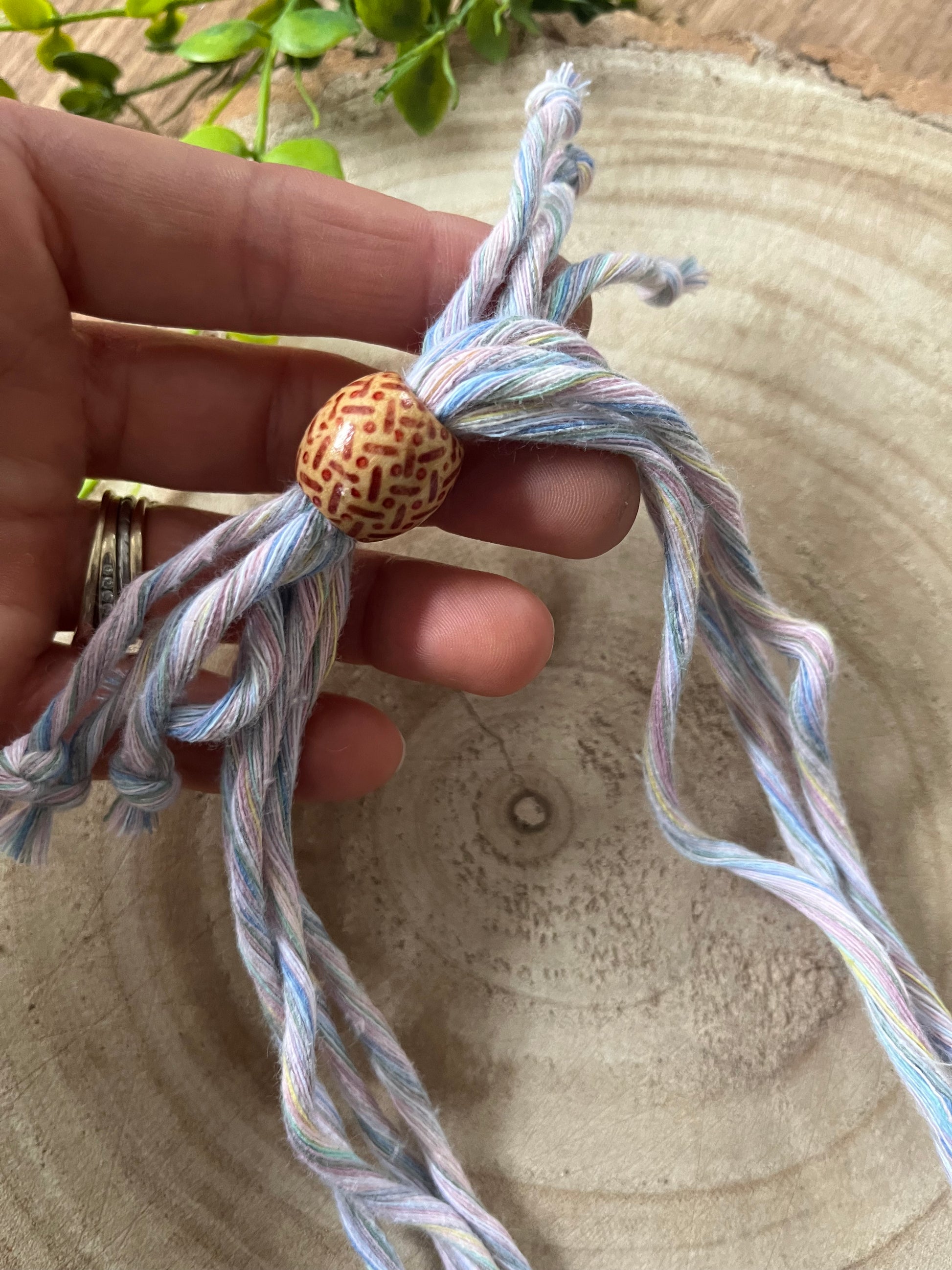 A hand holding a small patterned wooden bead on a crossbody phone strap made with pastel coloured yarn.