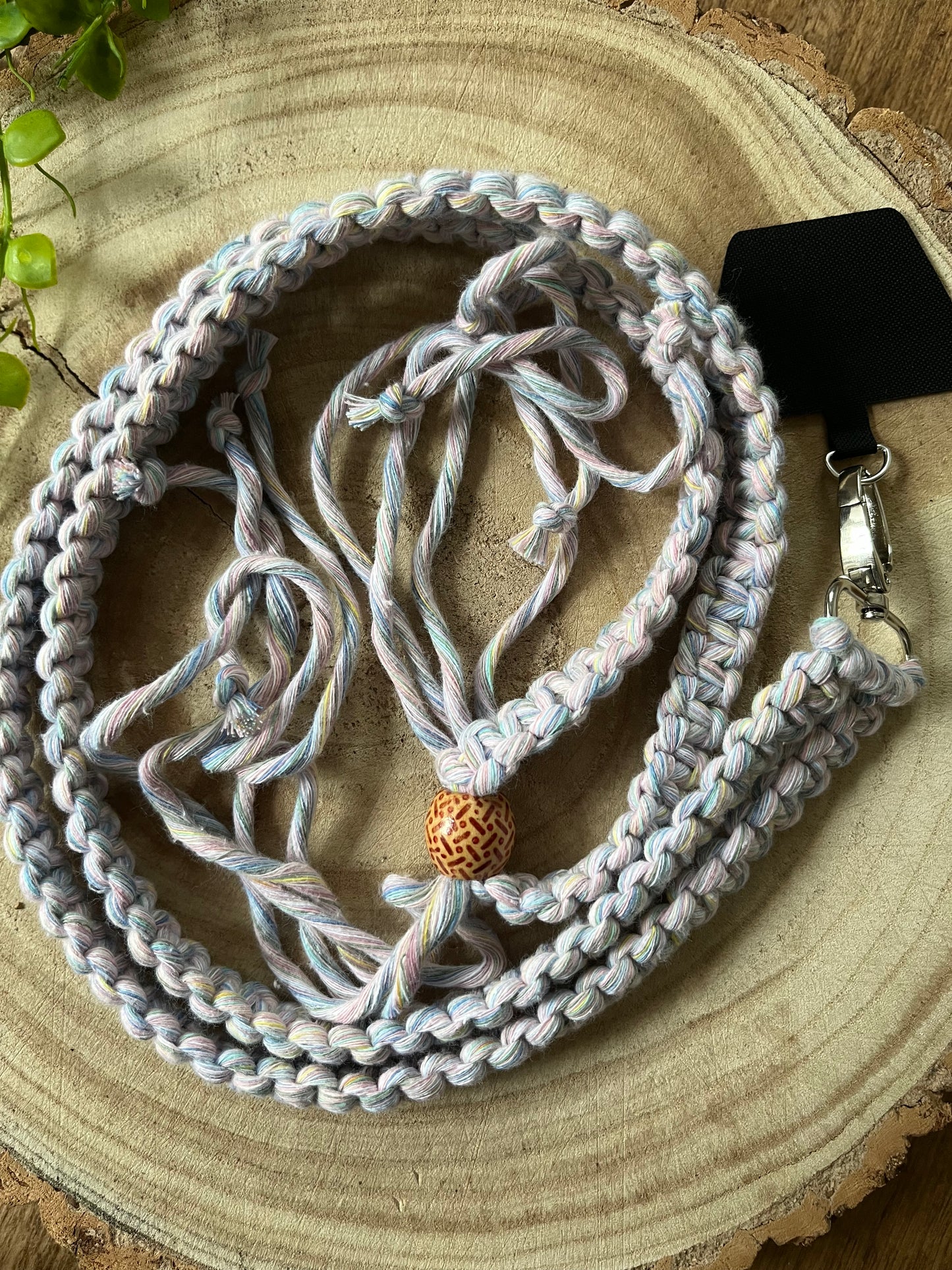 A long crossbody phone strap made with pastel yarn, a patterned wooden bead and metal clasp has a universal tether attached to it, and is coiled neatly on a round wooden surface.