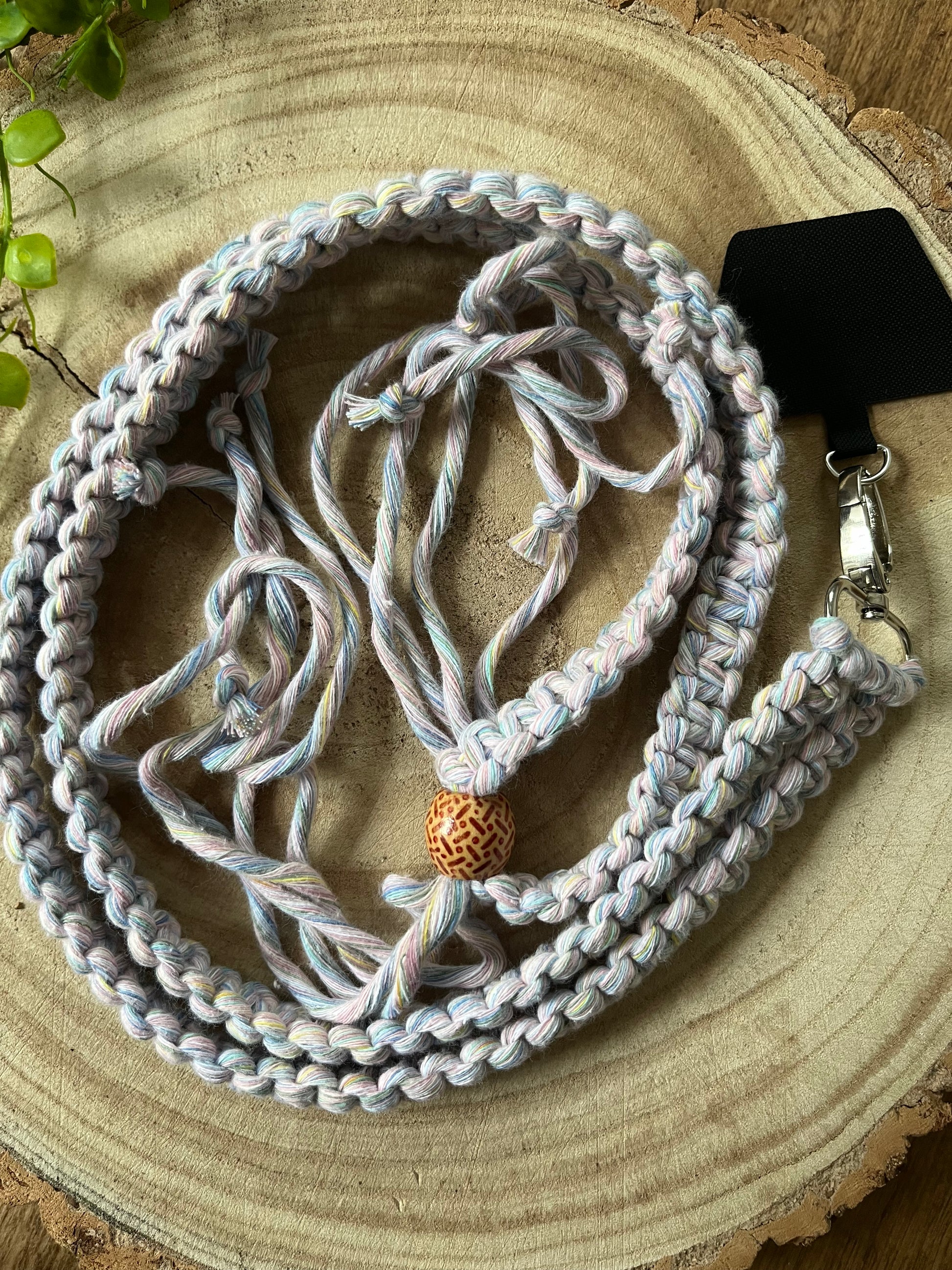 A long crossbody phone strap made with pastel yarn, a patterned wooden bead and metal clasp has a universal tether attached to it, and is coiled neatly on a round wooden surface.