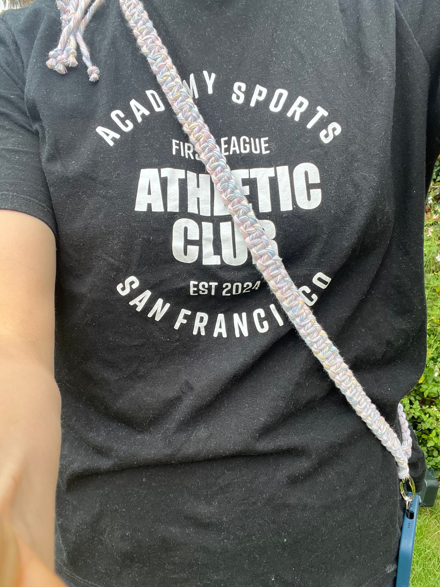 A woman stands outside wearing a black t shirt with white text on it, she has a crossbody phone strap across her body attached to a blue phone case.