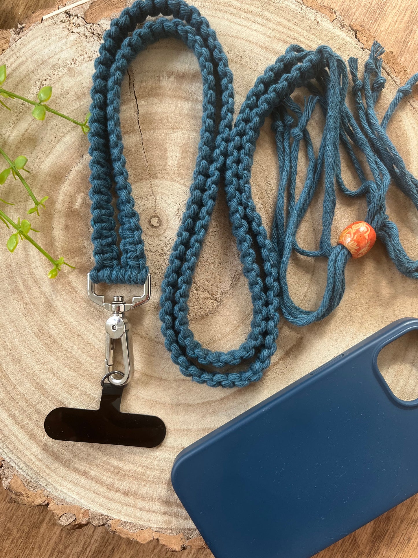 An adjustable crossbody phone strap with universal tether is coiled neatly on a wooden surface. The strap is made with petrol blue, recycled cotton with a metal clasp and a small wooden bead. An empty blue smartphone case and a small green plant are also visible in the scene.