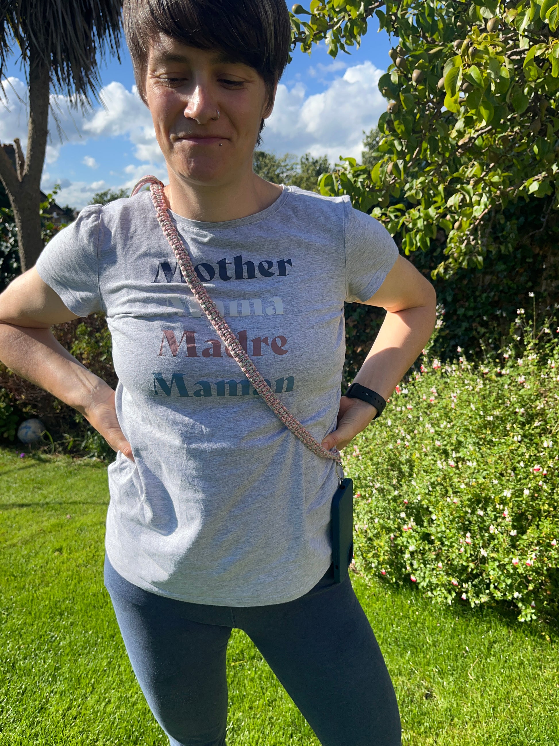 A woman stands outdoors in a sunny garden with her hands on her hips. She is wearing a grey t-shirt with the words "Mother," "Mama," "Madre," and "Maman" written in different colours, blue leggings, and an adjustable crossbody phone strap made with rainbow yarn. Lush greenery and a partly cloudy sky are in the background.