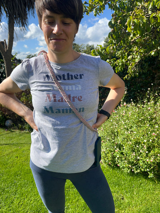 A woman stands outdoors in a sunny garden with her hands on her hips. She is wearing a grey t-shirt with the words "Mother," "Mama," "Madre," and "Maman" written in different colours, blue leggings, and an adjustable crossbody phone strap made with rainbow yarn. Lush greenery and a partly cloudy sky are in the background.