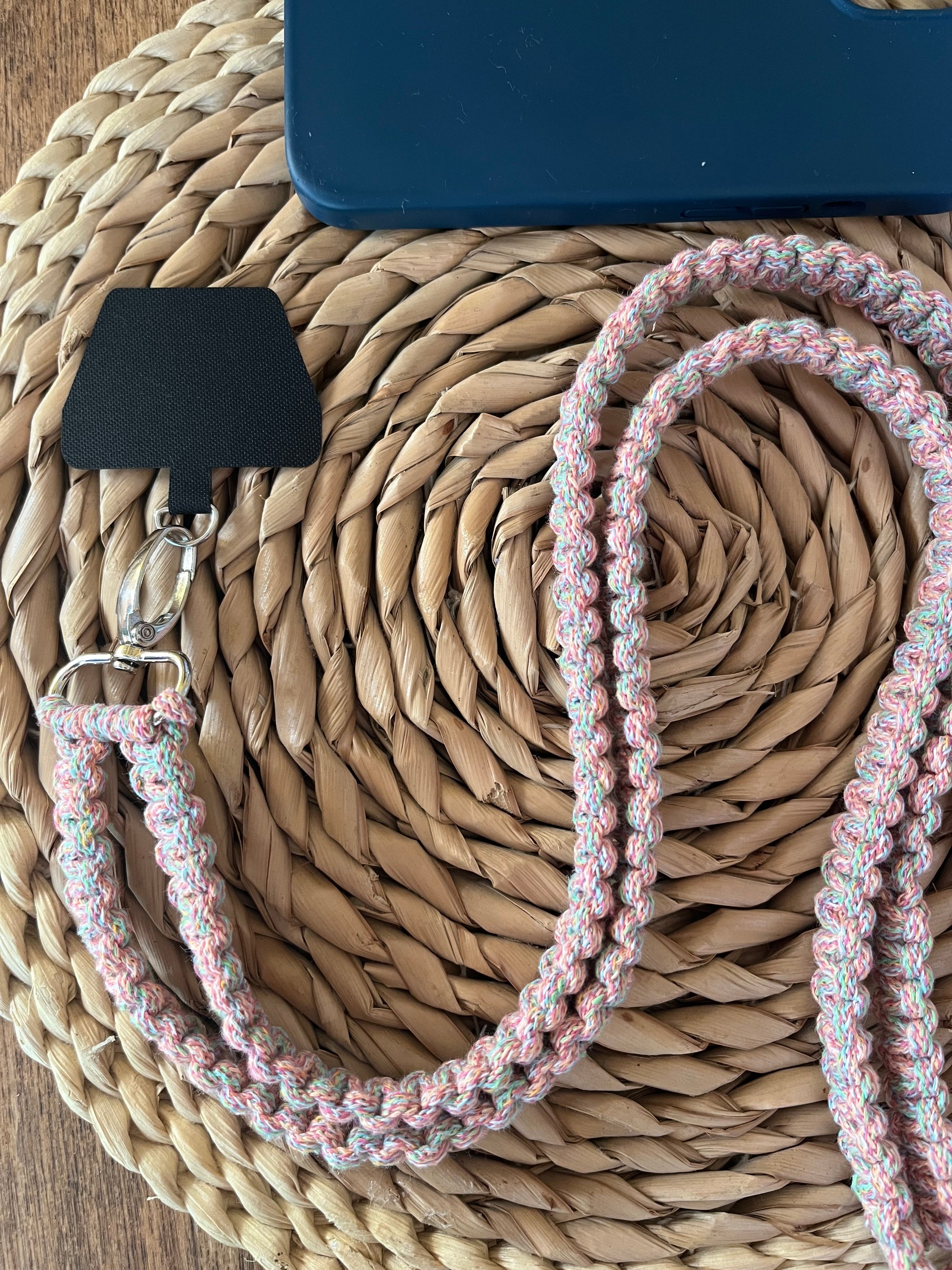 A woven, multicoloured adjustable crossbody phone strap with universal tether and a metal clasp on a circular, woven placemat.