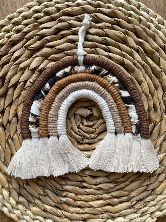 A macrame rainbow made in different shades of brown, plus an arch of animal print yarn, with fluffy white tassels, is laid flat on a woven mat