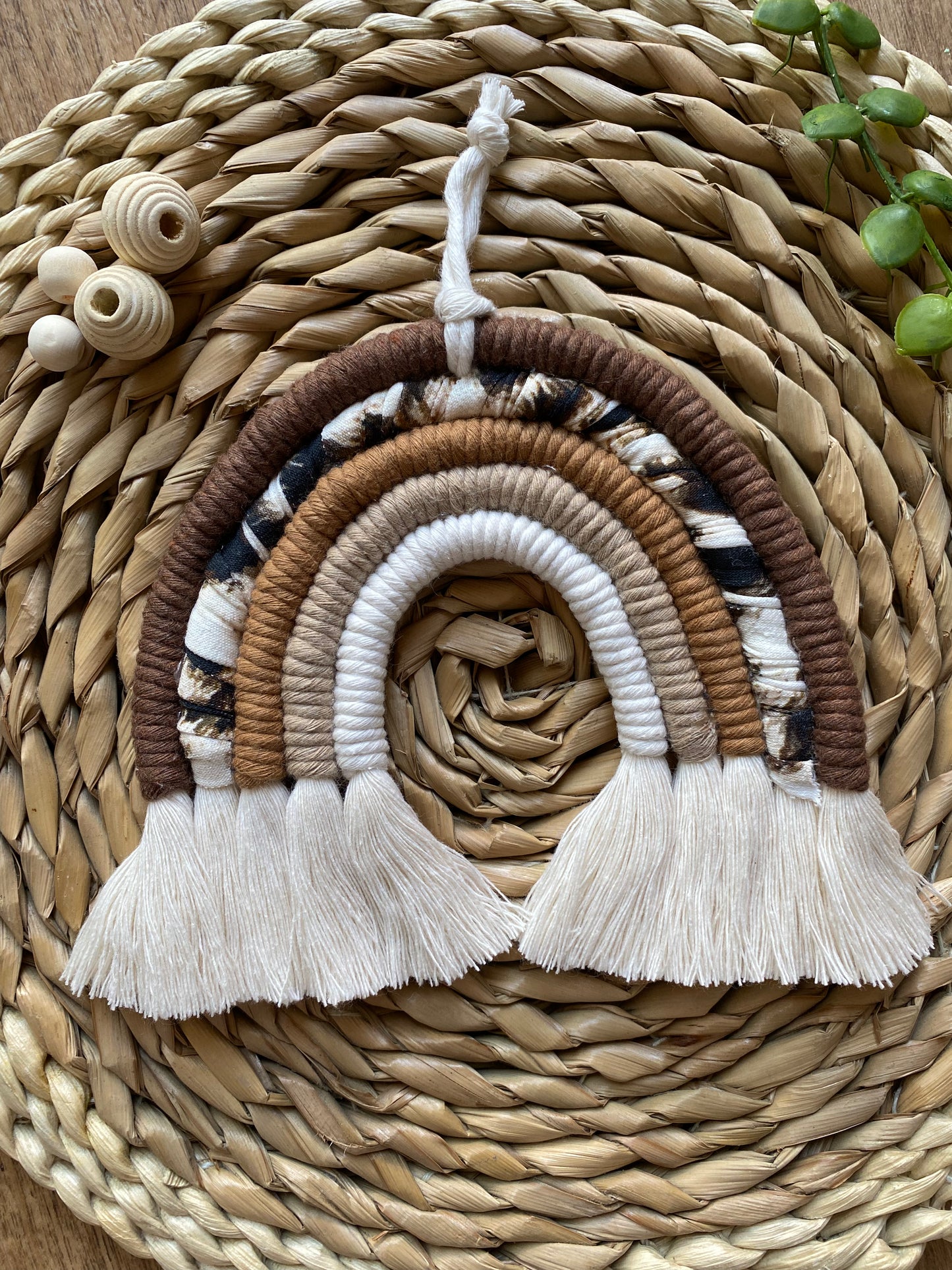 A macrame rainbow made in different shades of brown, with an arch of animal print yarn and fluffy white tassels, is laid flat on a woven mat alongside some wooden beads and green plant vines.