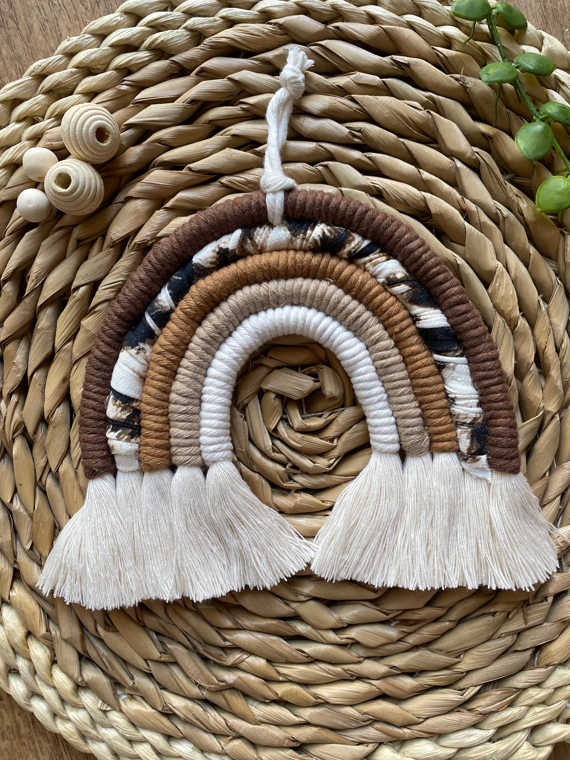 A macrame rainbow made in different shades of brown, with an arch of animal print yarn and fluffy white tassels, is laid flat on a woven mat alongside some wooden beads and green plant vines.