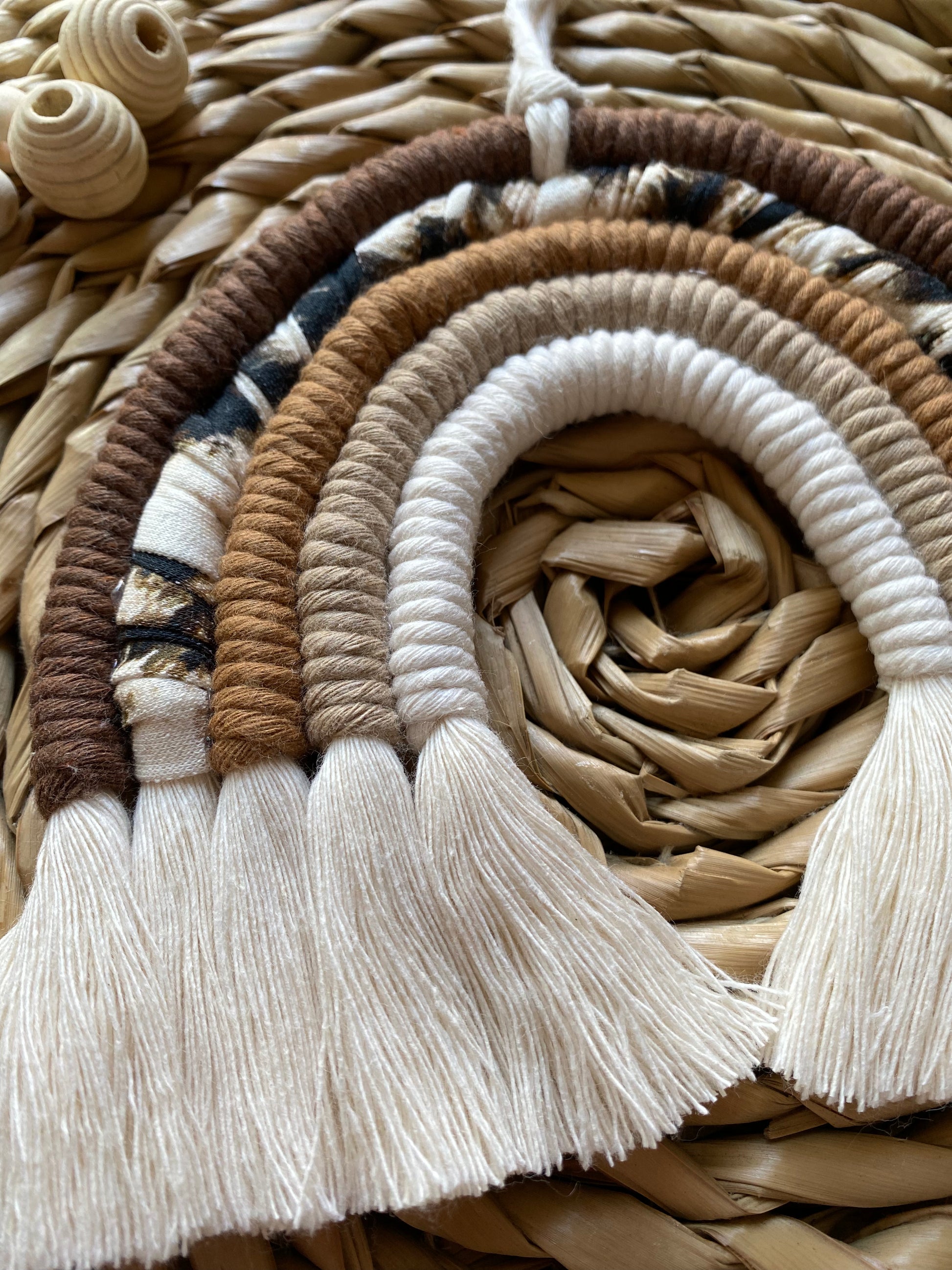Close up of a macrame rainbow made in different shades of brown, with an arch of animal print yarn and fluffy white tassels, on a woven mat alongside some wooden beads.