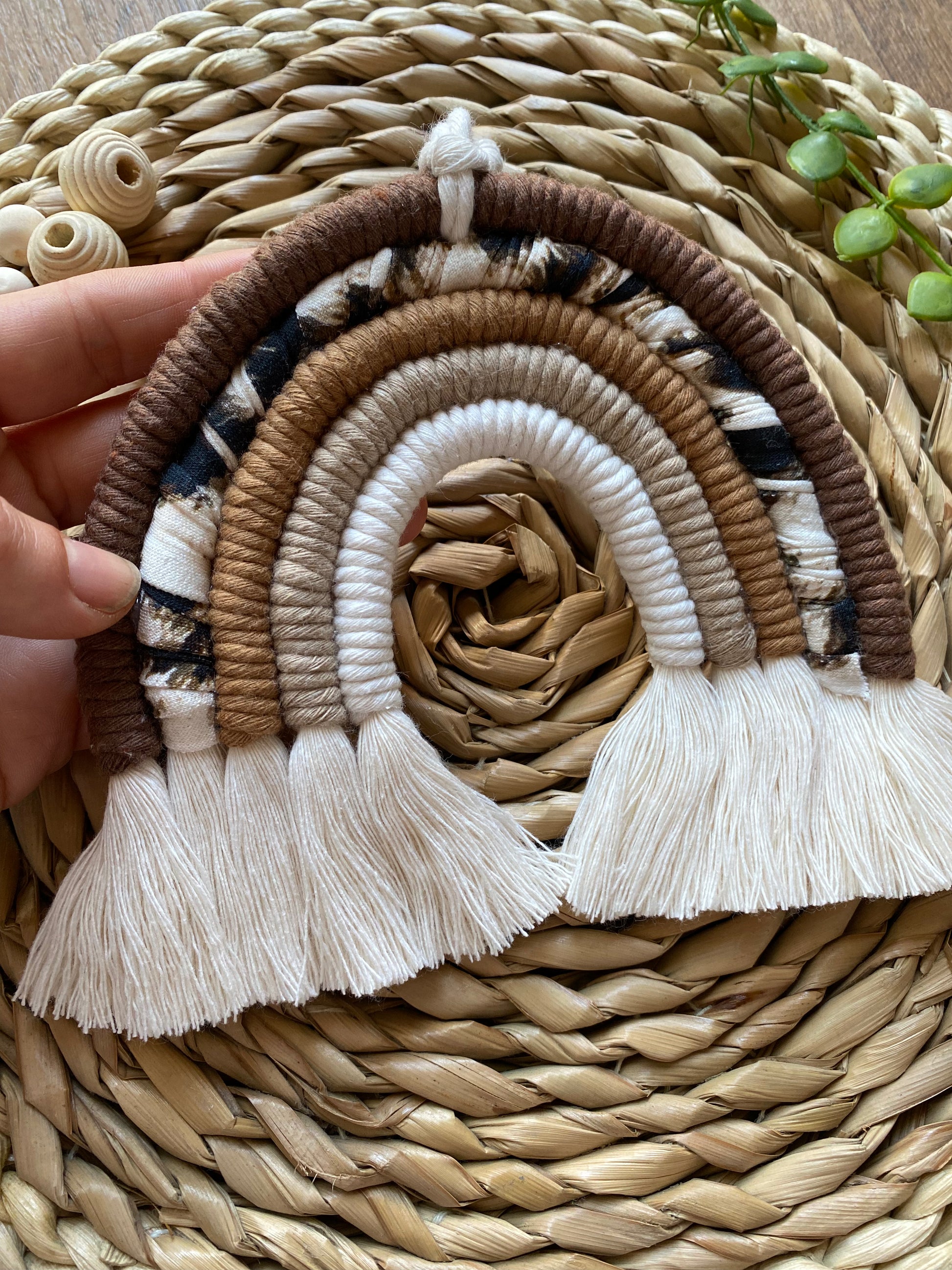 A hand holds a macrame rainbow made in different shades of brown, with an arch of animal print yarn and fluffy white tassels, on a woven mat alongside some wooden beads and green plant vines.