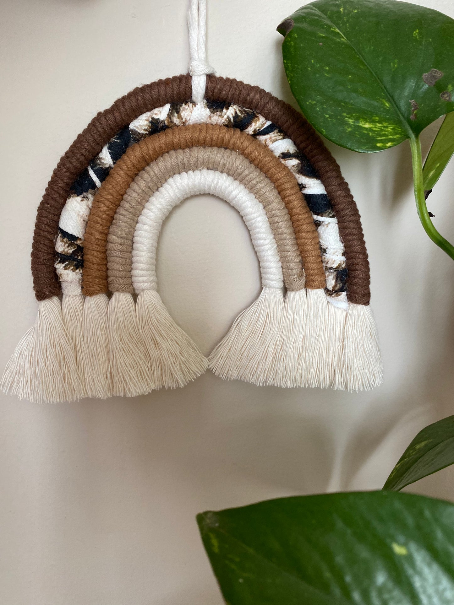 A macrame rainbow made in different shades of brown, with an arch of animal print yarn and fluffy white tassels, is hanging against a beige wall, next to a large green houseplant.