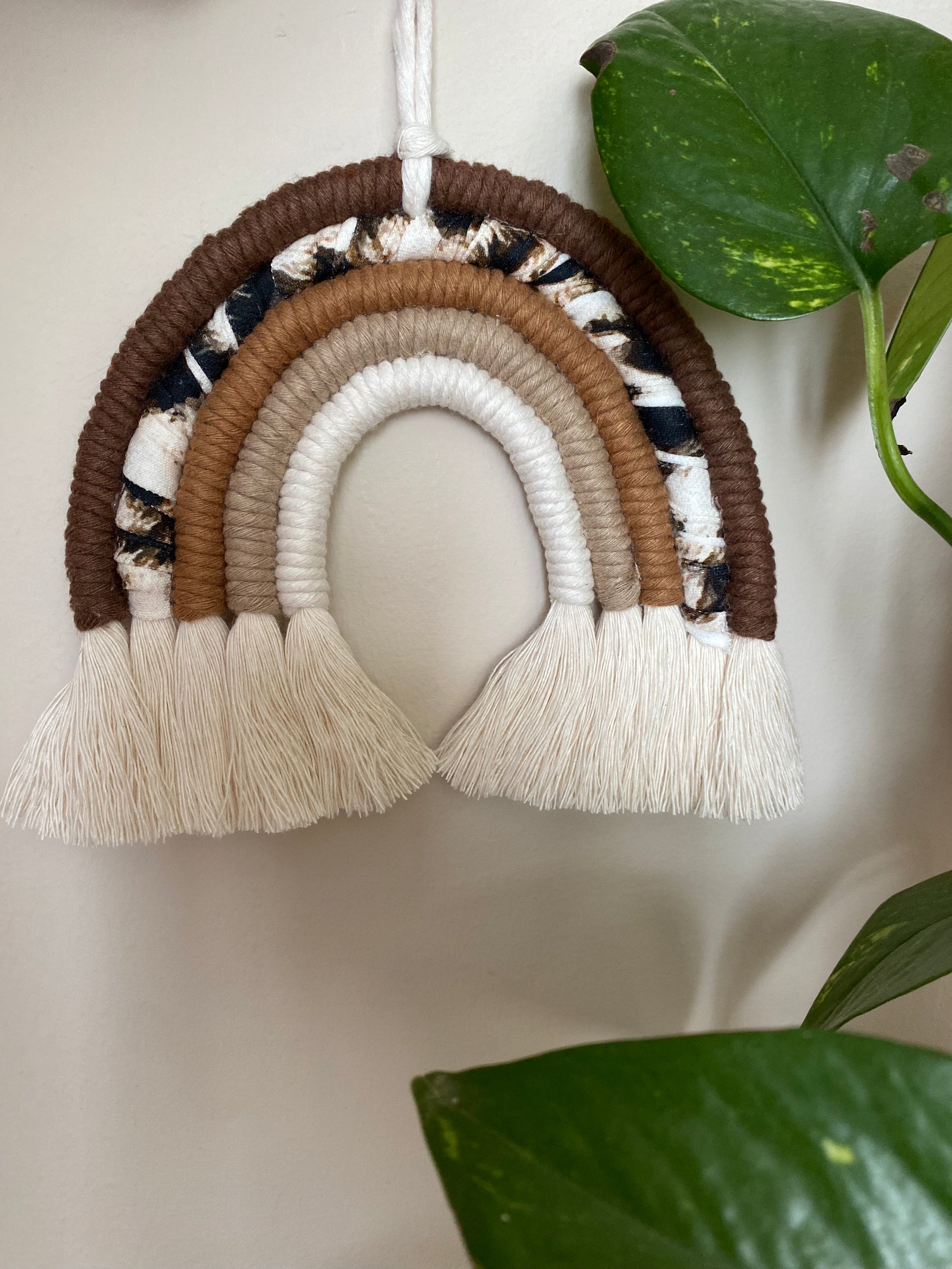 A macrame rainbow made in different shades of brown, with an arch of animal print yarn and fluffy white tassels, is hanging against a beige wall, next to a large green houseplant.