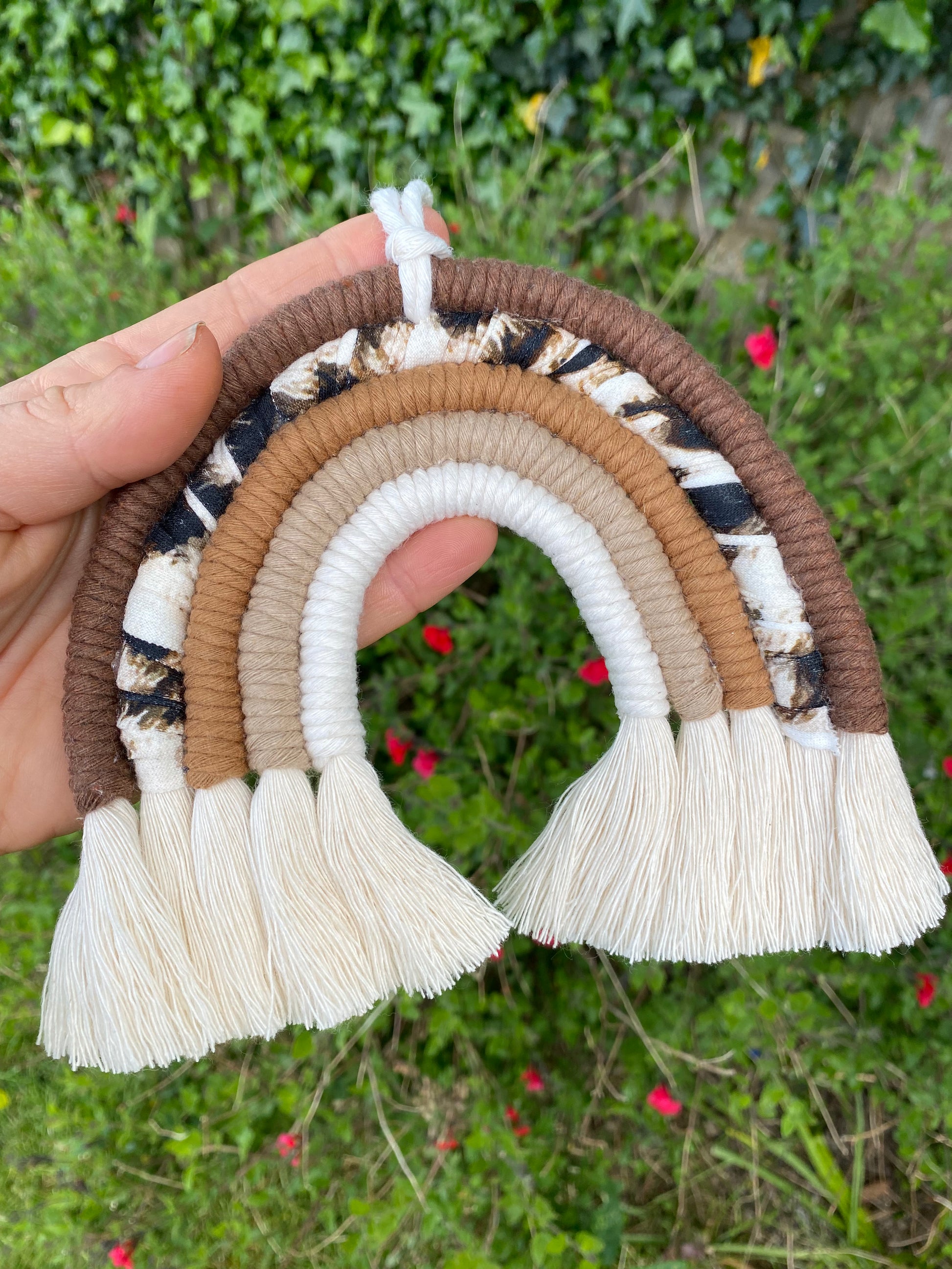 A hand is holding a macrame rainbow made in different shades of brown, with an arch of animal print yarn and fluffy white tassels, with green and pink plants in the background.