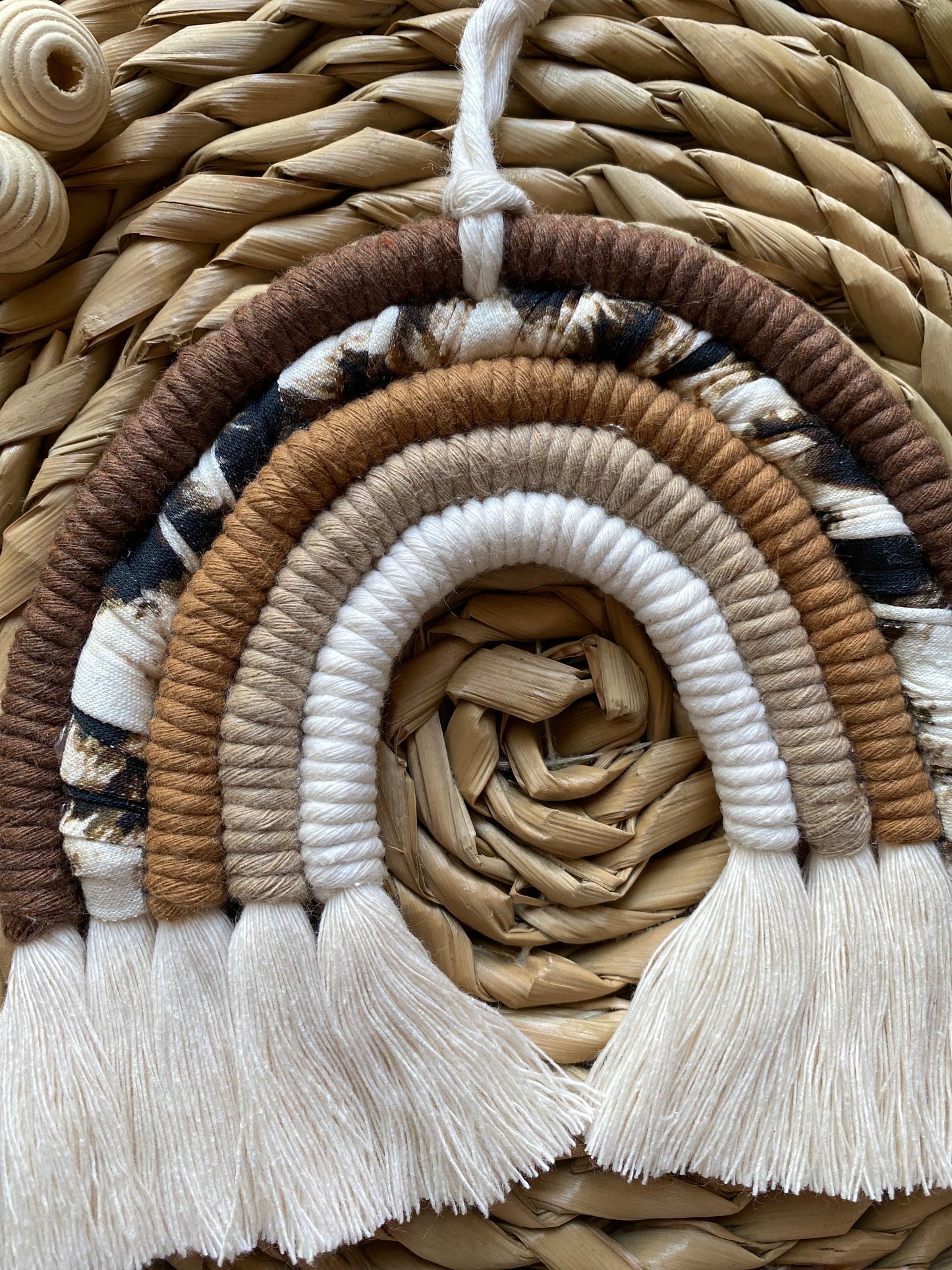 A macrame rainbow made in different shades of brown, with an arch of animal print yarn and fluffy white tassels, is laid flat on a woven mat alongside some wooden beads.