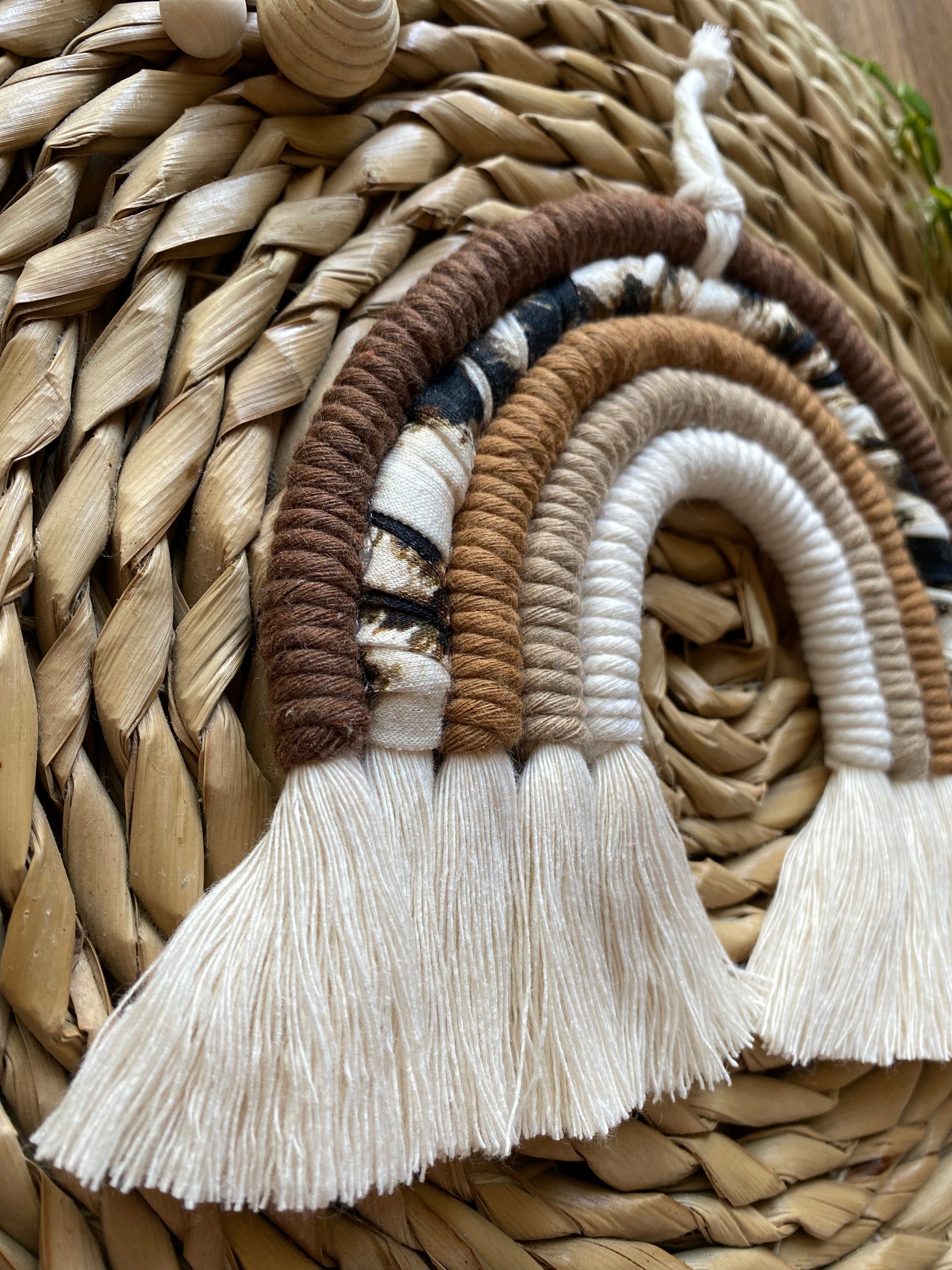 A close up side angled view of a macrame rainbow made in different shades of brown, with an arch of animal print yarn and fluffy white tassels.