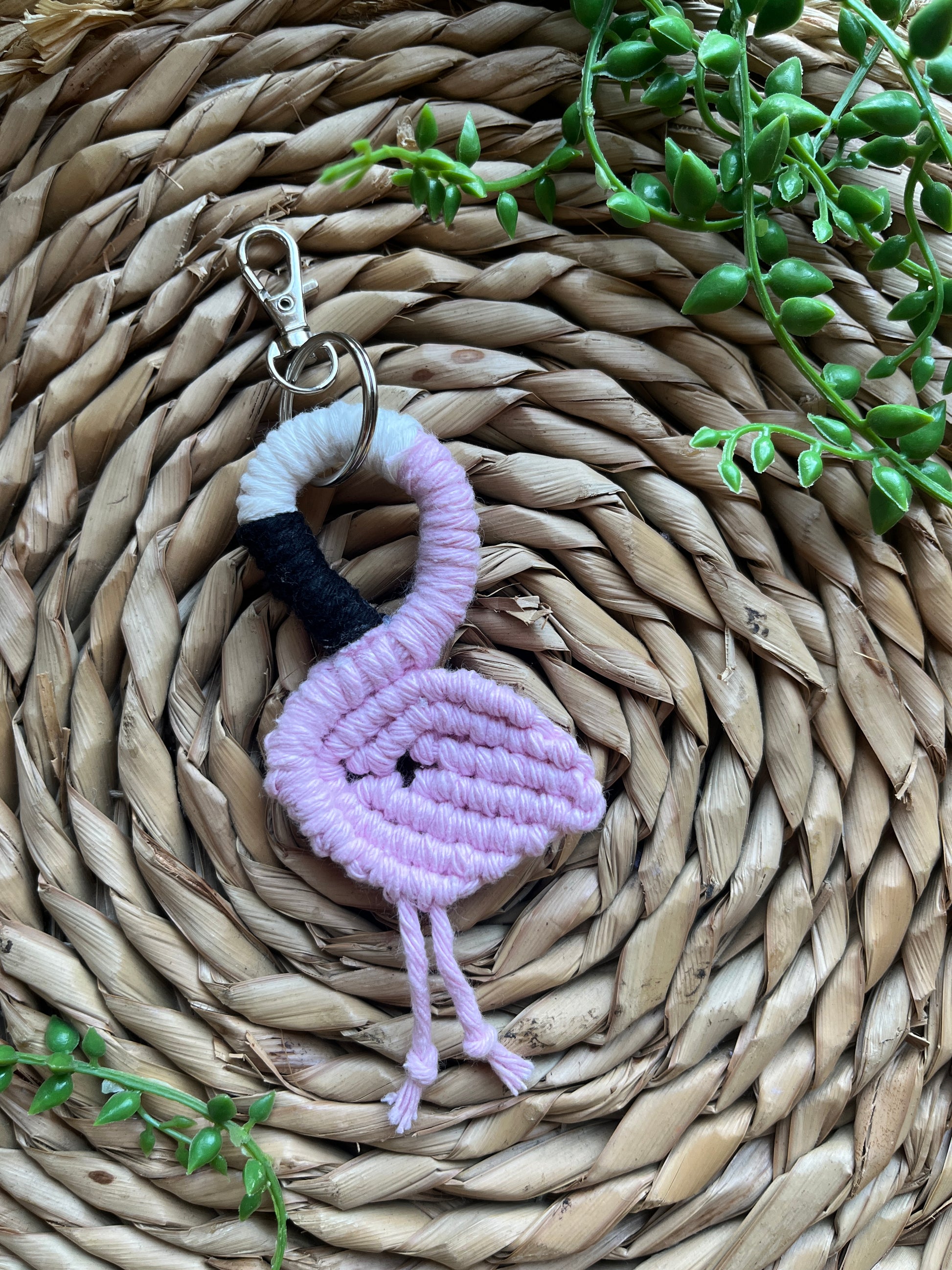Handmade macrame keychain in the shape of a flamingo, made using light pink recycled cotton, on a metal split ring with lobster clasp. The keyring is displayed on a round wicker surface next to some green plant vines.