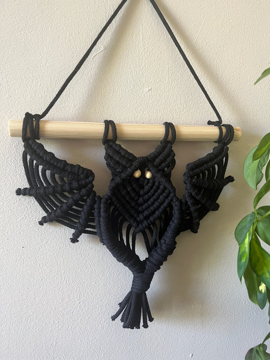 Black macrame bat wall hanging with wooden bead eyes on a wooden dowel, hanging against a beige painted wall next to a green houseplant